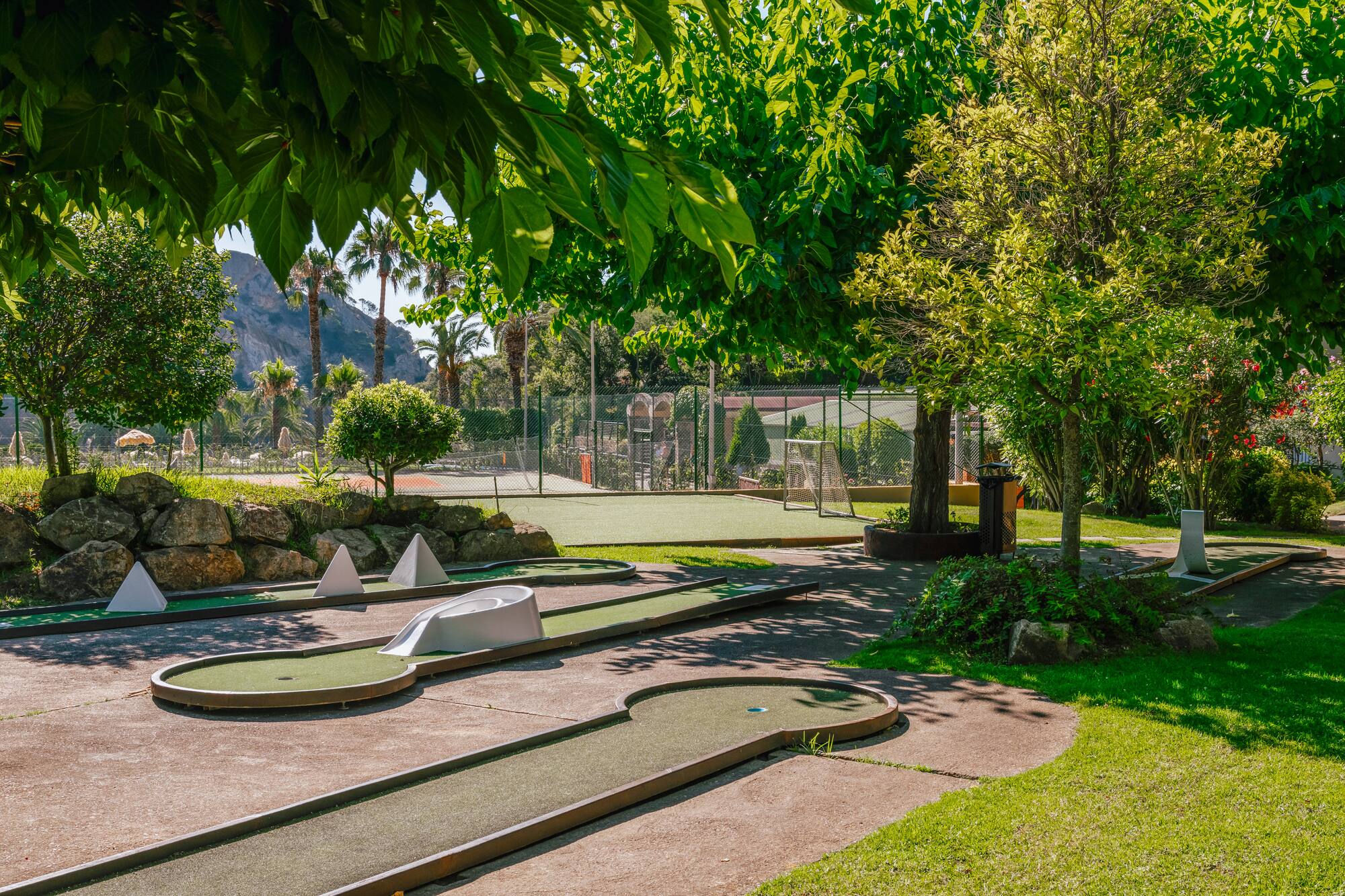 Outdoor mini-golf course with green turf, lush trees, and mountain backdrop.