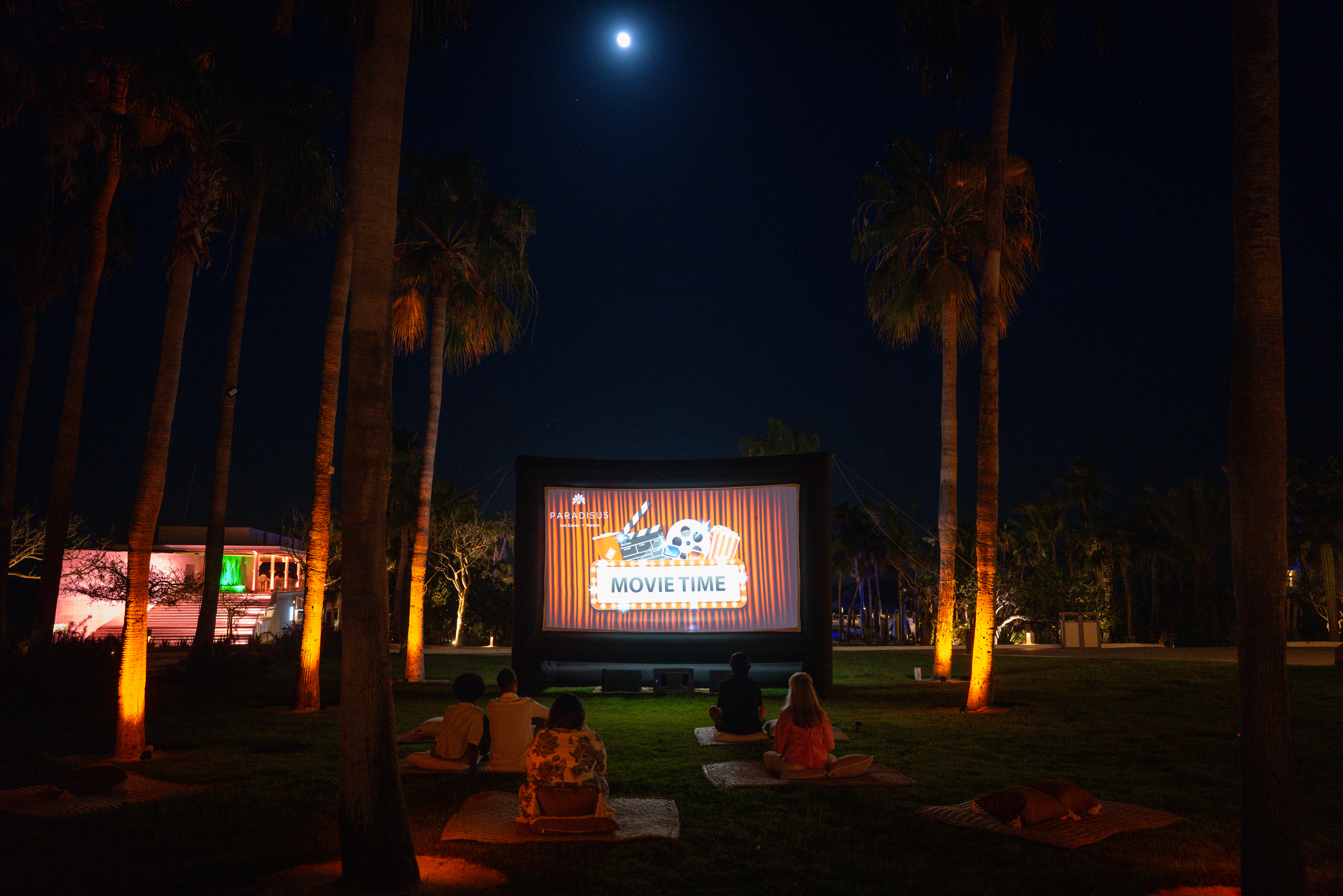 Outdoor movie night with audience watching large screen, palm trees, moon.