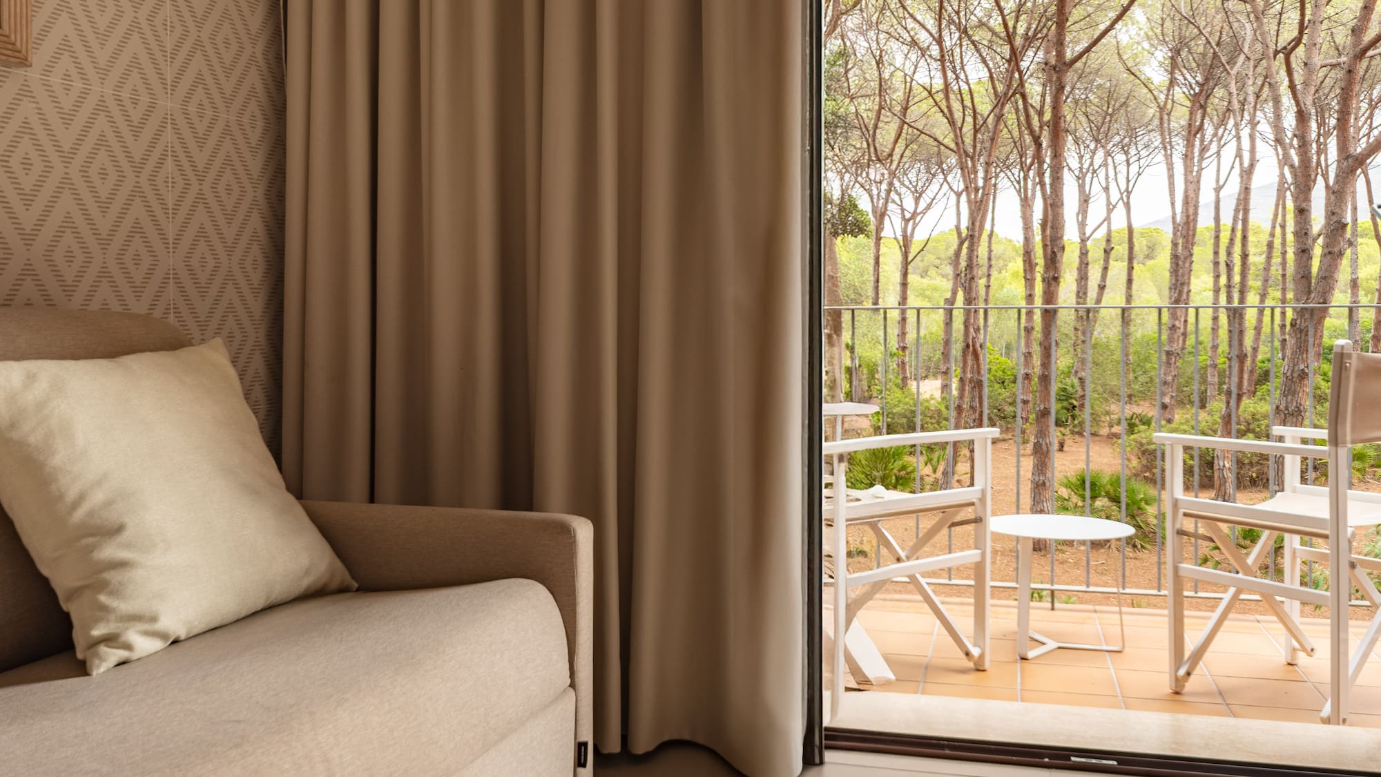 Hotel room with sofa, open balcony with outdoor seating and forest view.