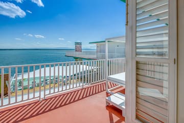 a deck with a white railing and a white building with a body of water in the background