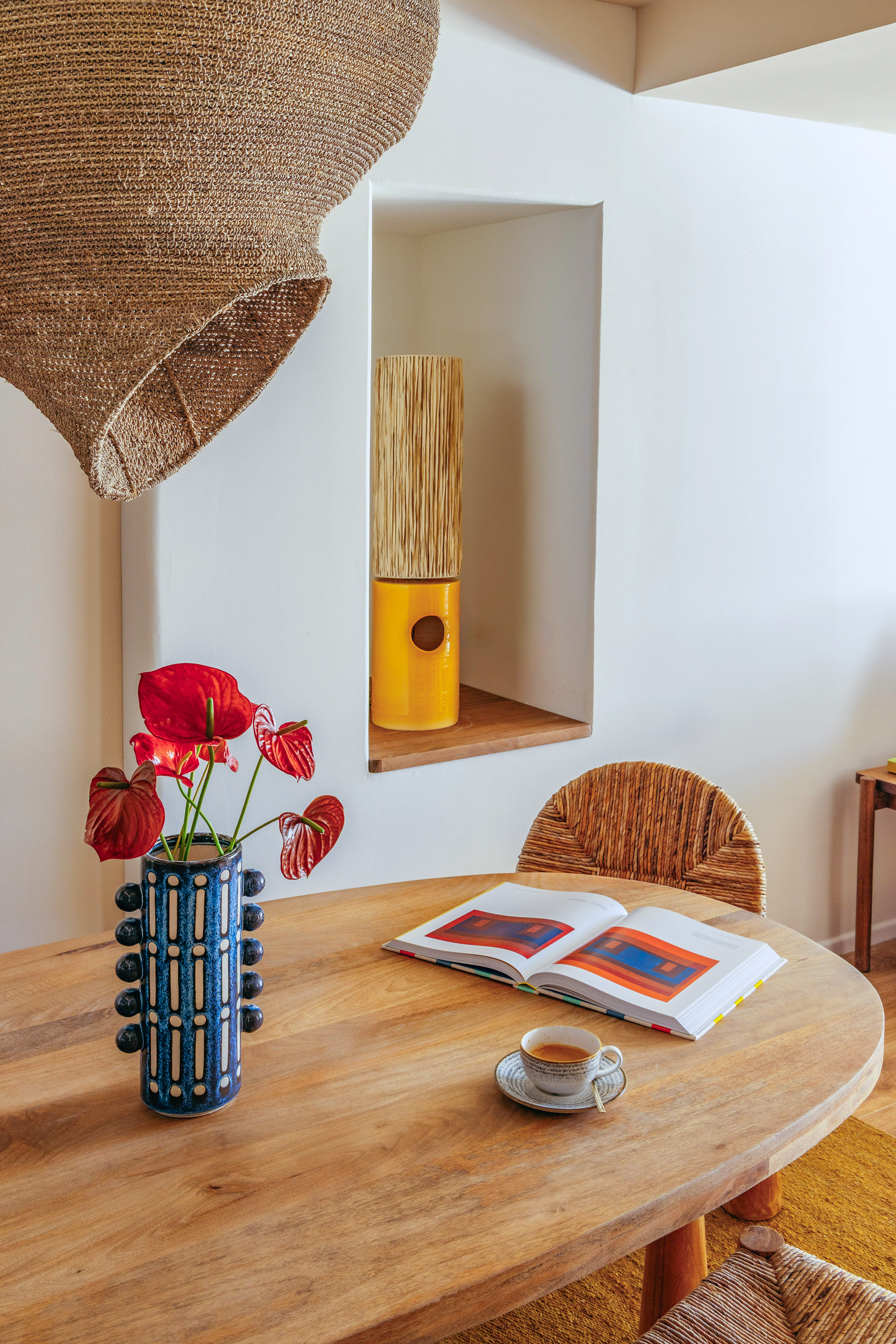 Round wood table with red anthurium vase, art book, coffee, woven chair and lamp.
