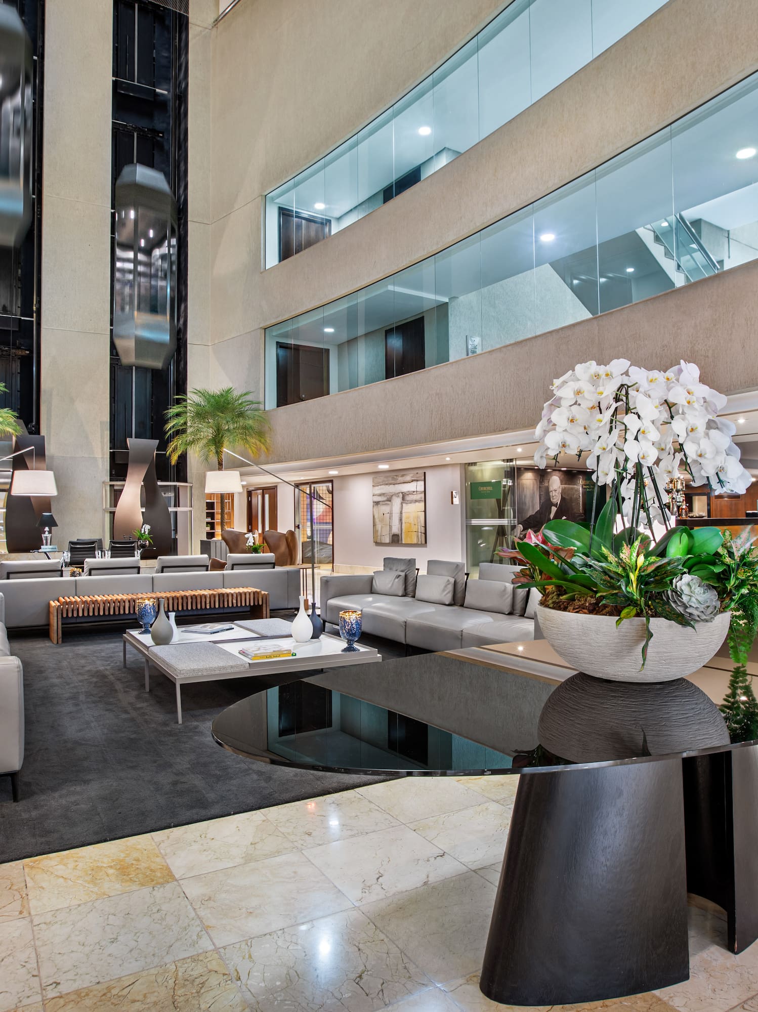 Spacious hotel lobby with modern lounge seating, glass elevators, and orchids.