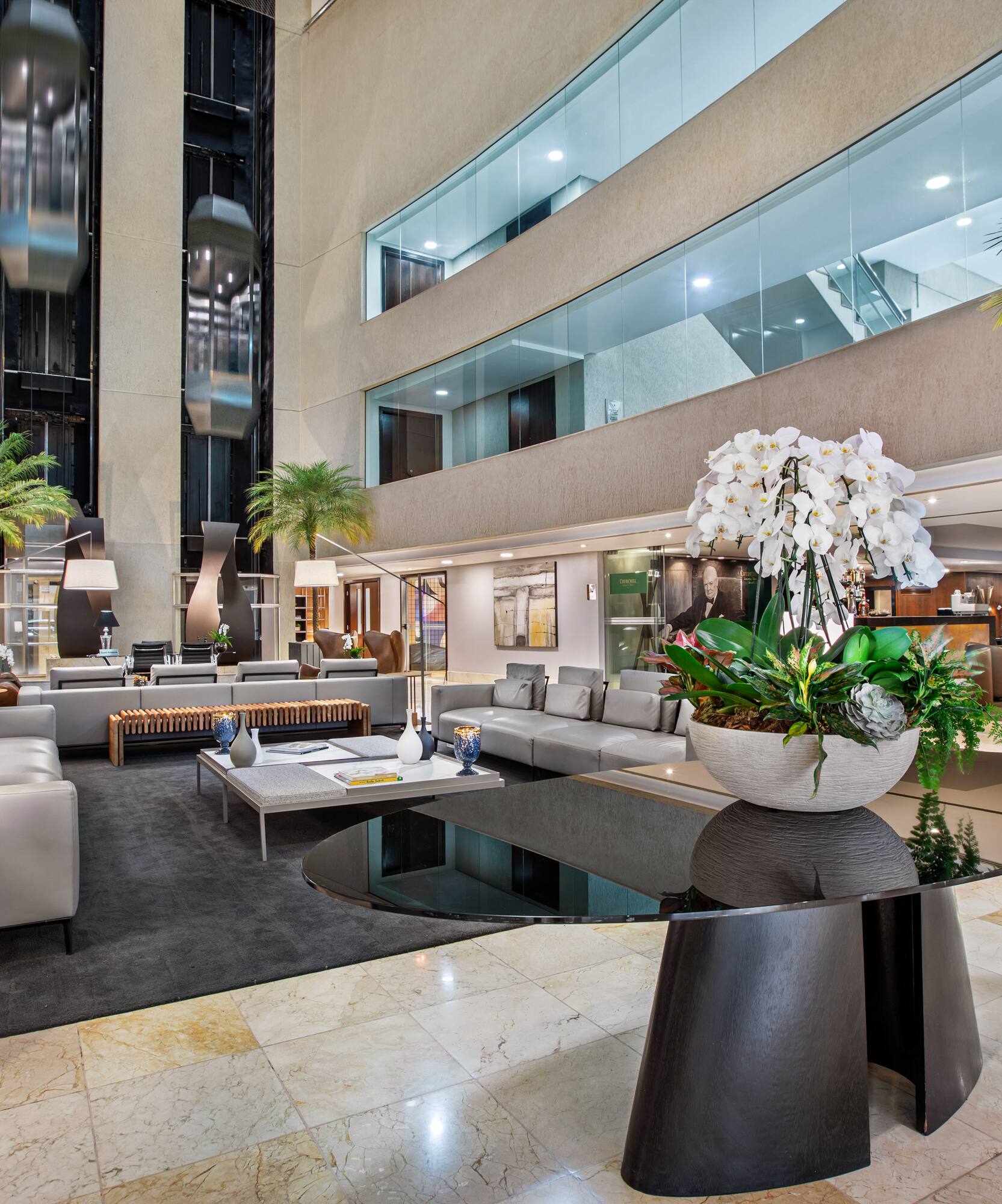Spacious hotel lobby with modern lounge seating, glass elevators, and orchids.