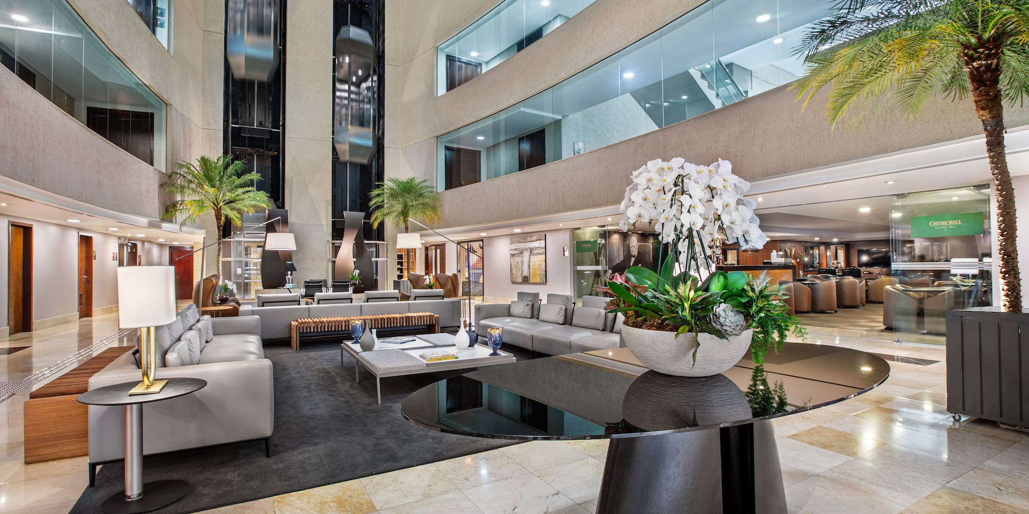 Modern hotel lobby with glass elevators, lounge seating, palm trees, and orchids.