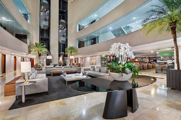 Modern hotel lobby with glass elevators, lounge seating, palm trees, and orchids.