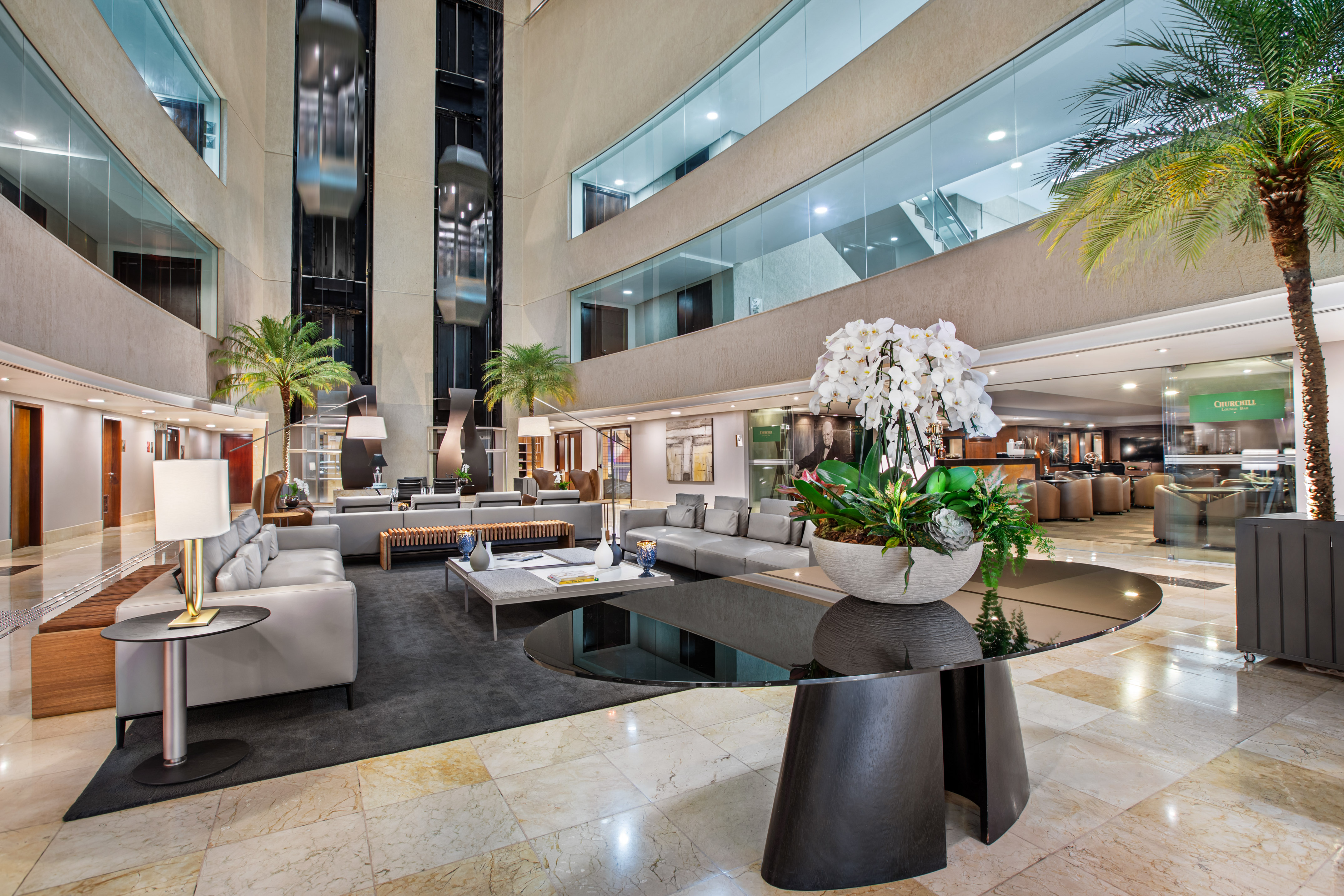 Spacious hotel lobby with modern lounge seating, glass elevators, and orchids.