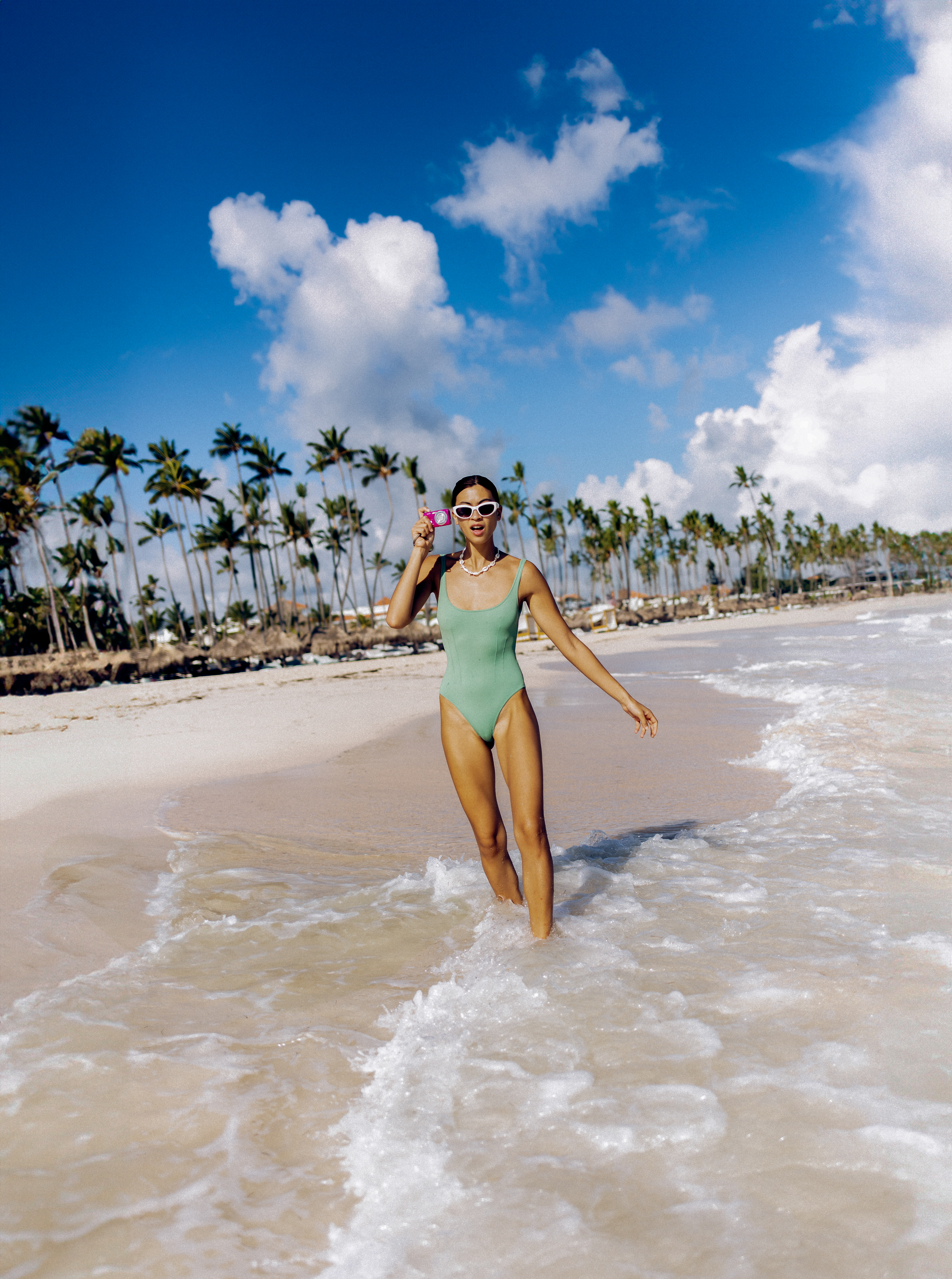 Woman in green swimsuit, sunglasses, walking in ocean waves on tropical beach.