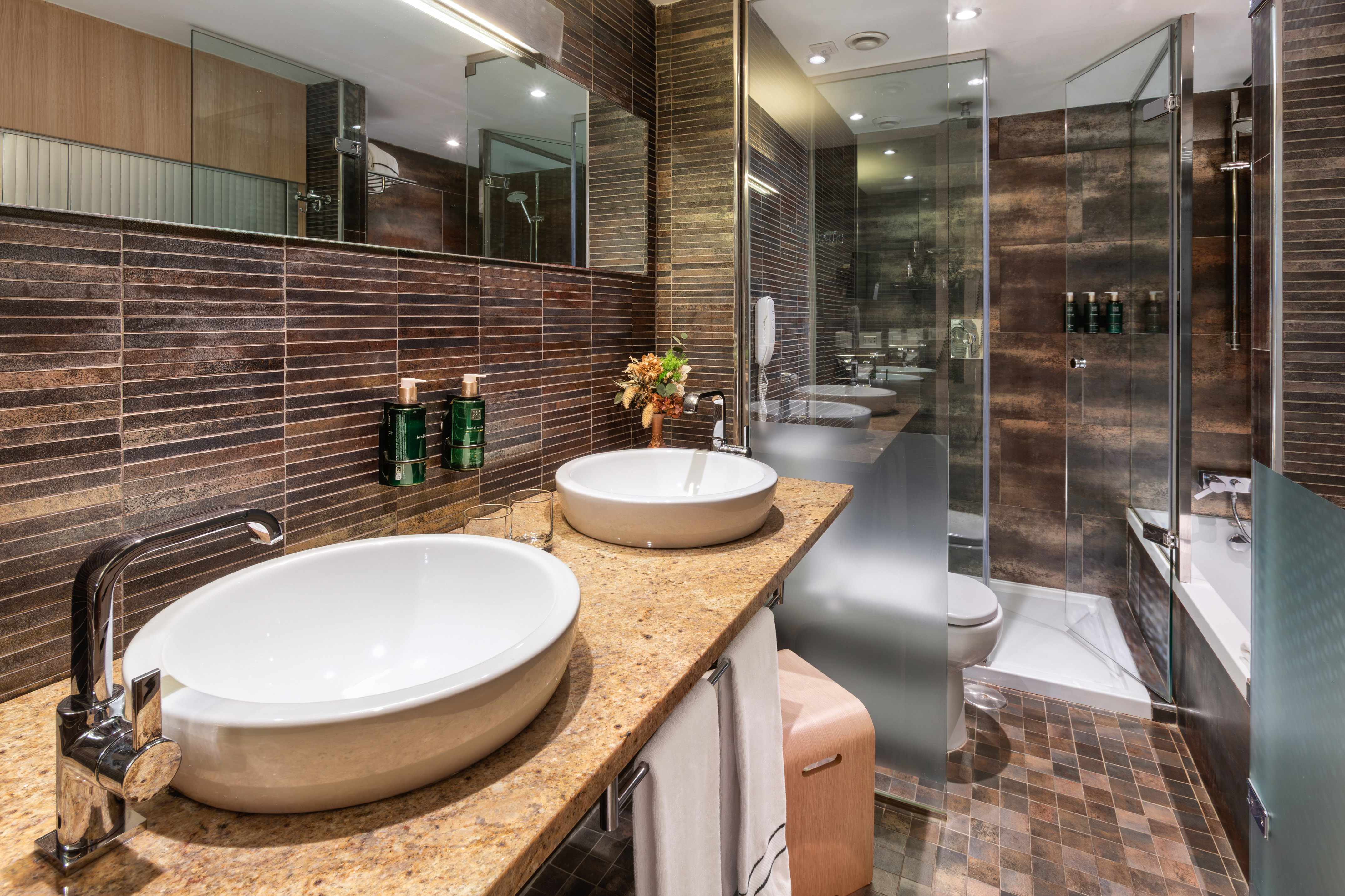 Luxury hotel bathroom with double sinks, brown tiles, and glass shower.