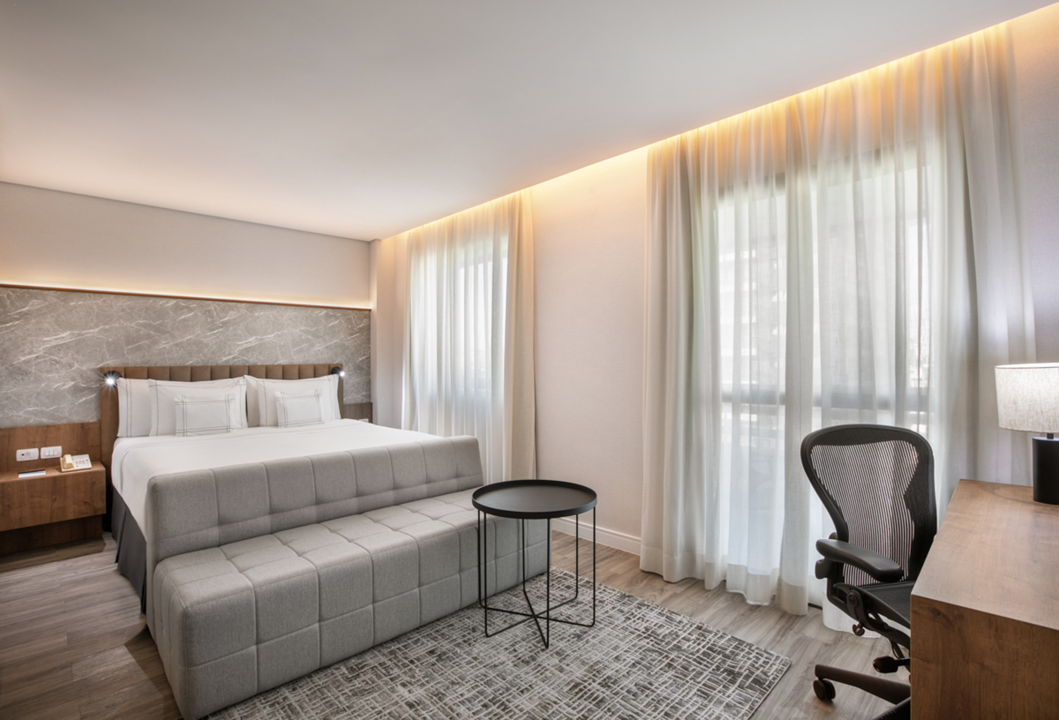 Modern hotel bedroom with king bed, gray bench, work desk, and ergonomic chair.