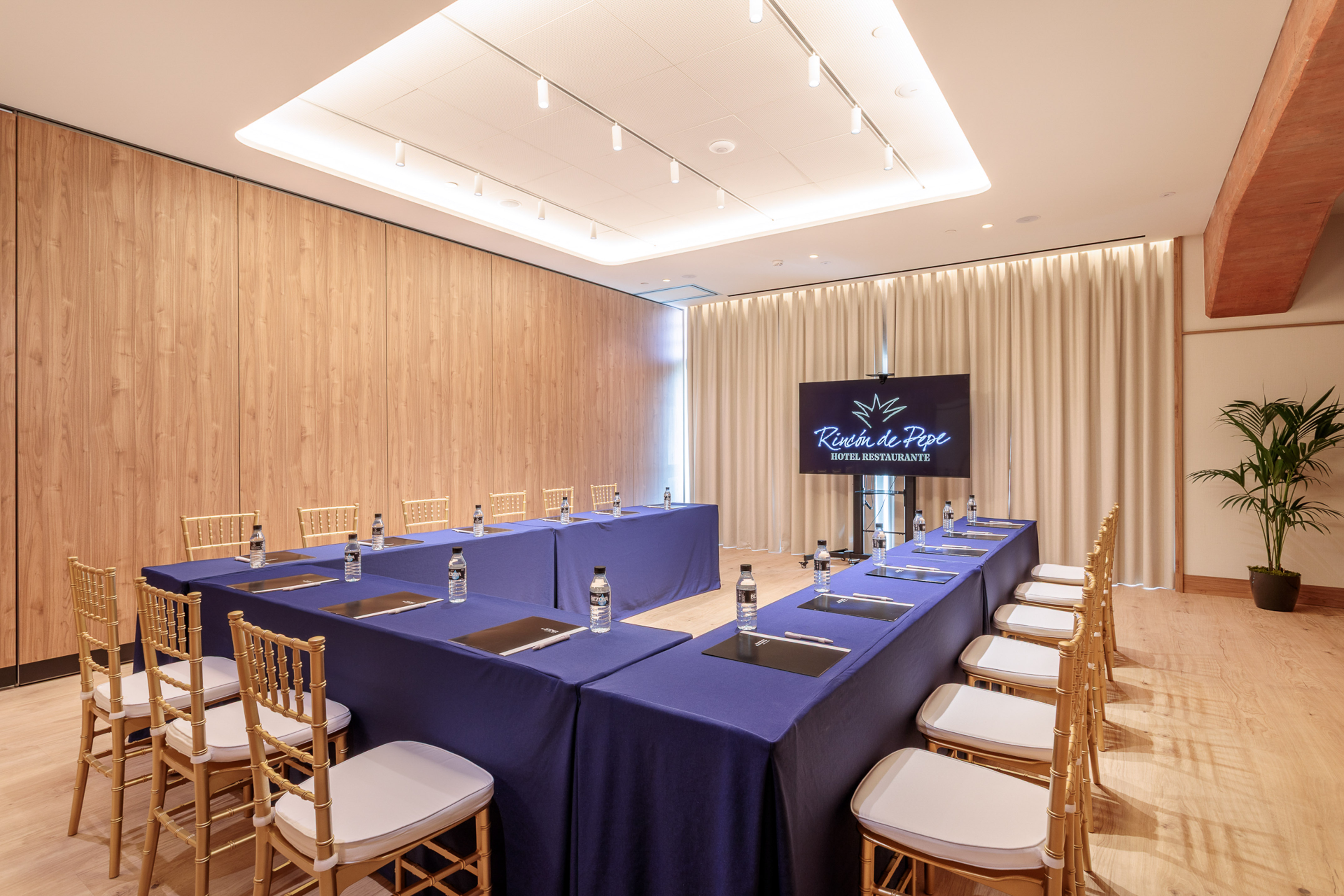 Rincón de Pepe hotel meeting room with U-shaped tables, gold chairs, wood walls.