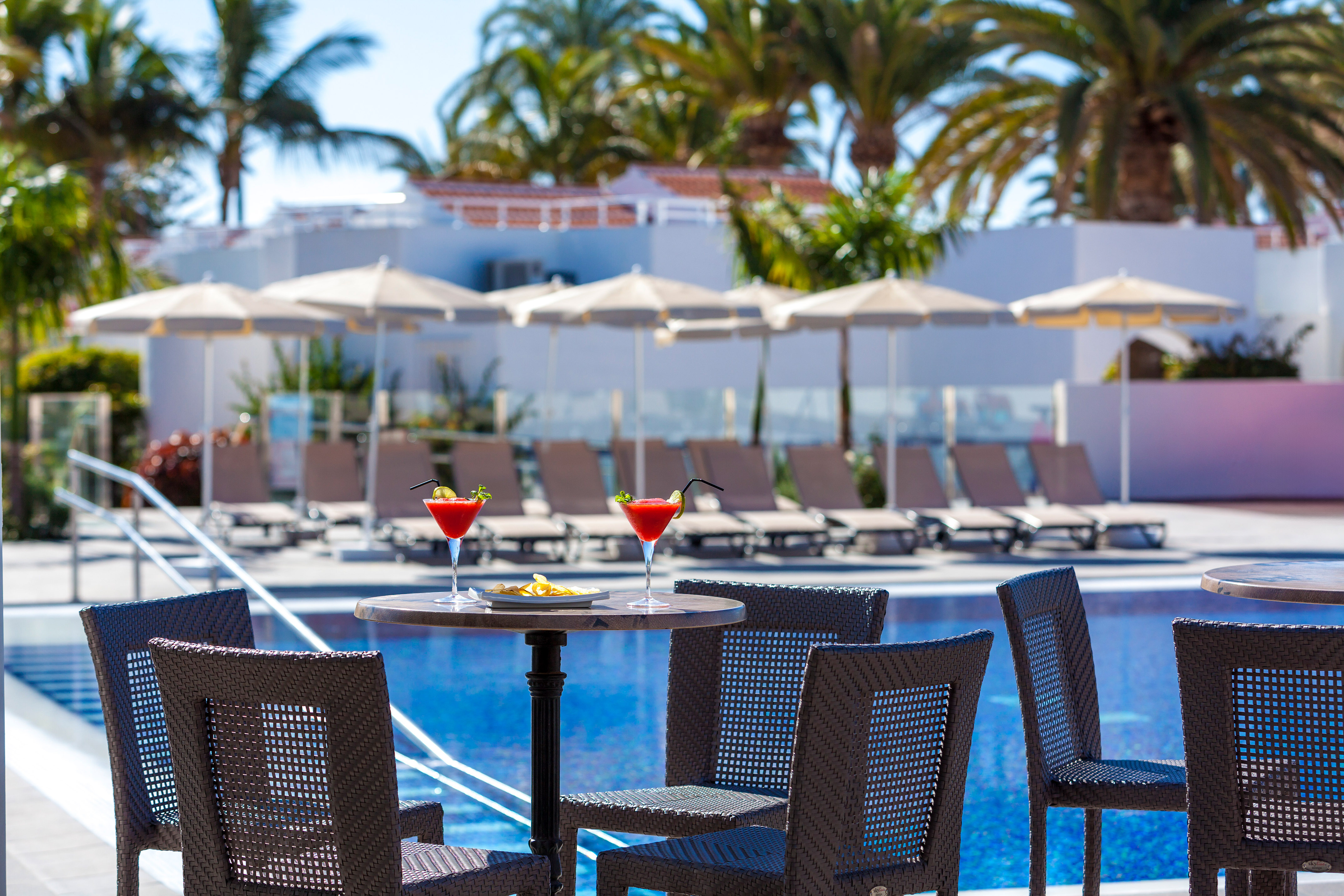Two red cocktails on a table by a sunny resort pool with palm trees.