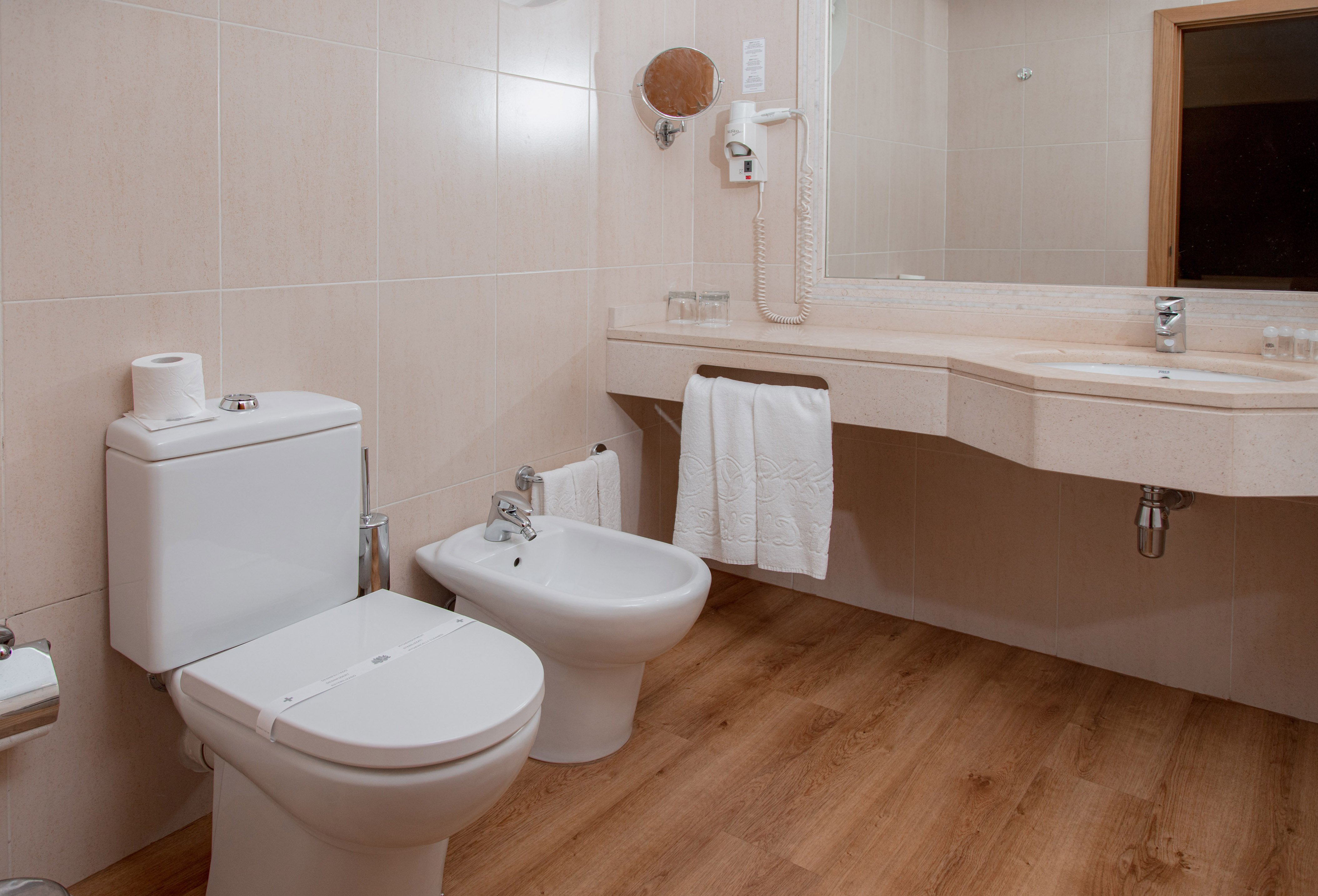 Bright, clean hotel bathroom with toilet, bidet, sink, mirror, and light wood floor.