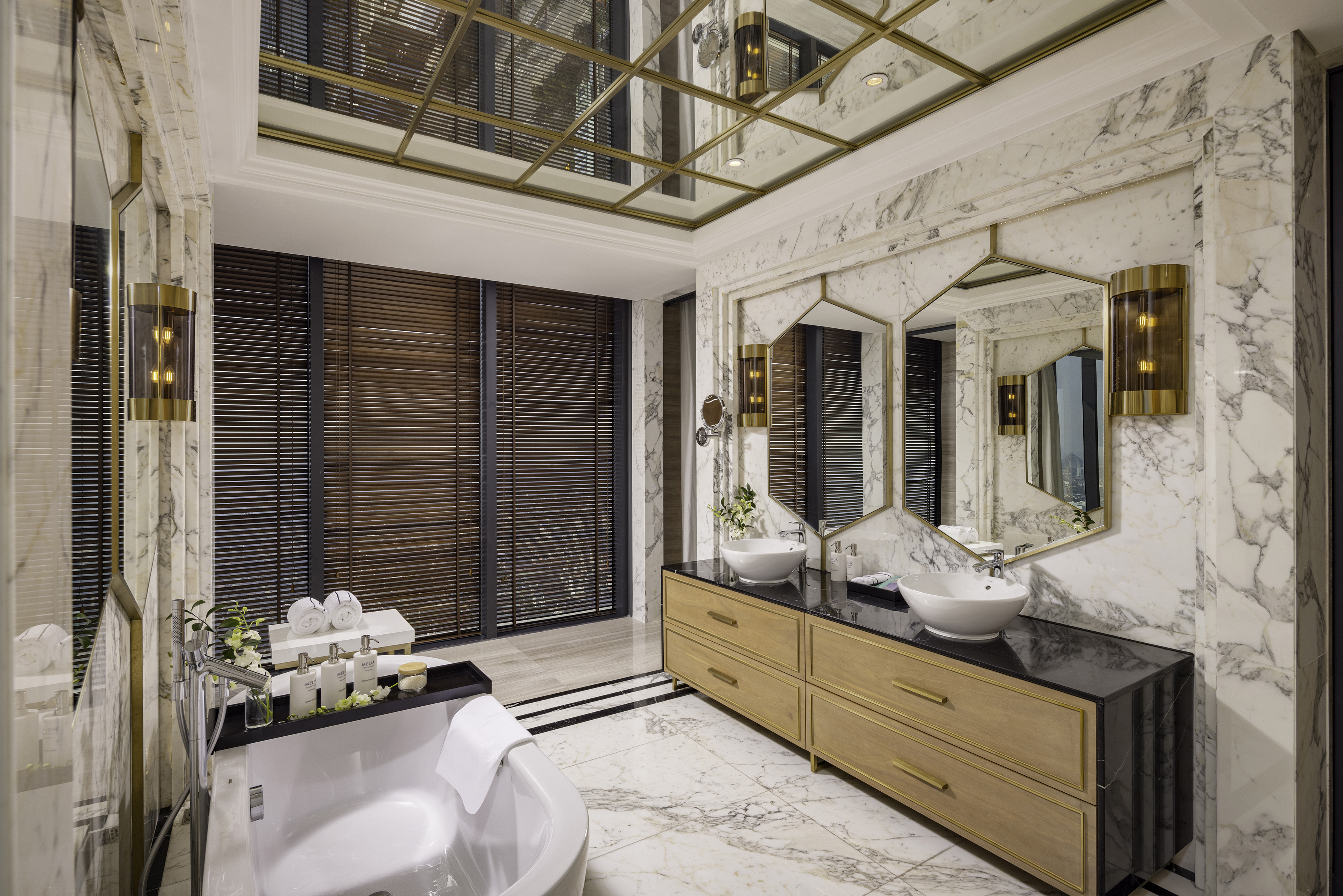 Luxury marble bathroom with bathtub, double vanity, gold accents, and wood blinds.
