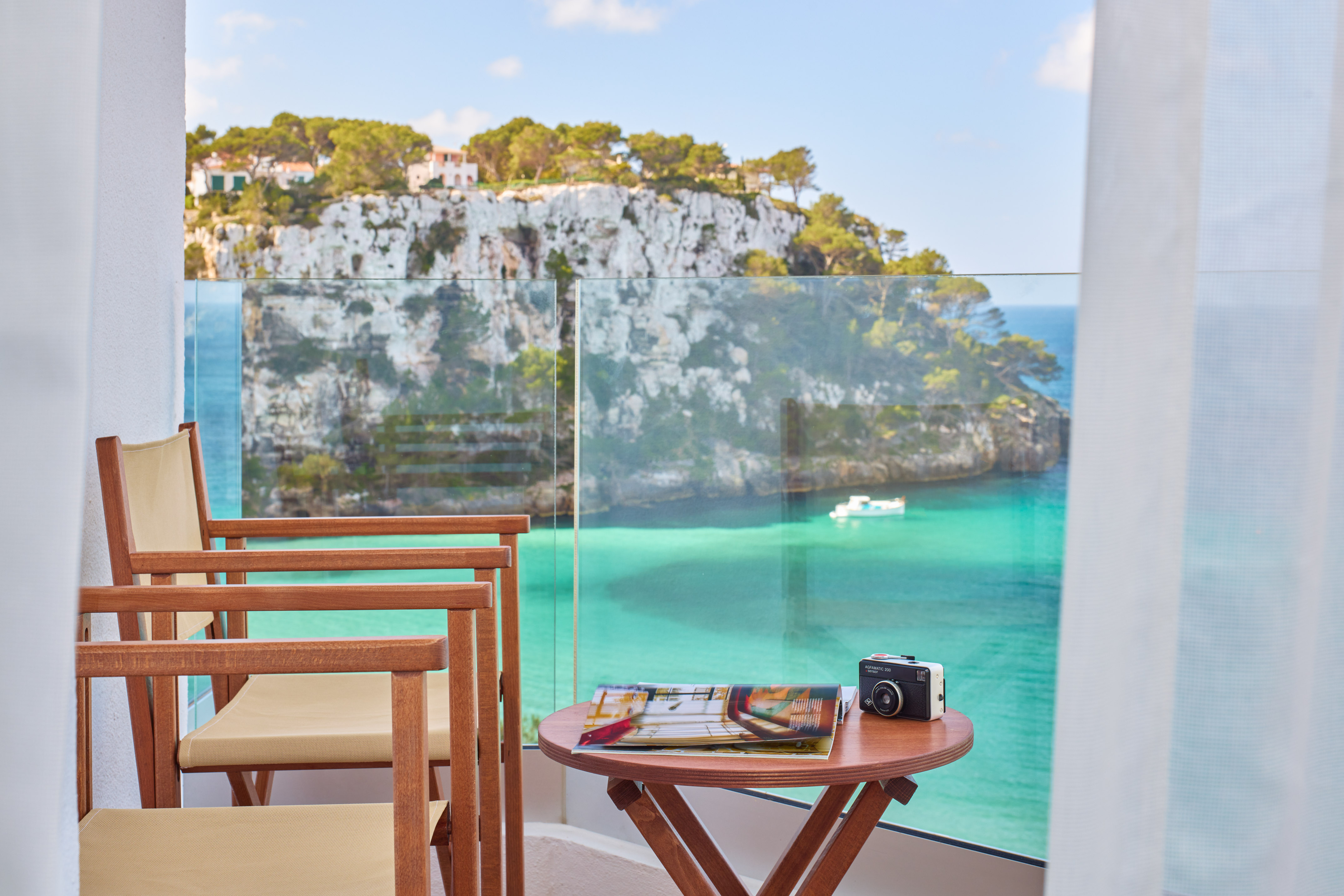 Hotel balcony with turquoise sea, coastal cliffs, chairs, and vintage camera.