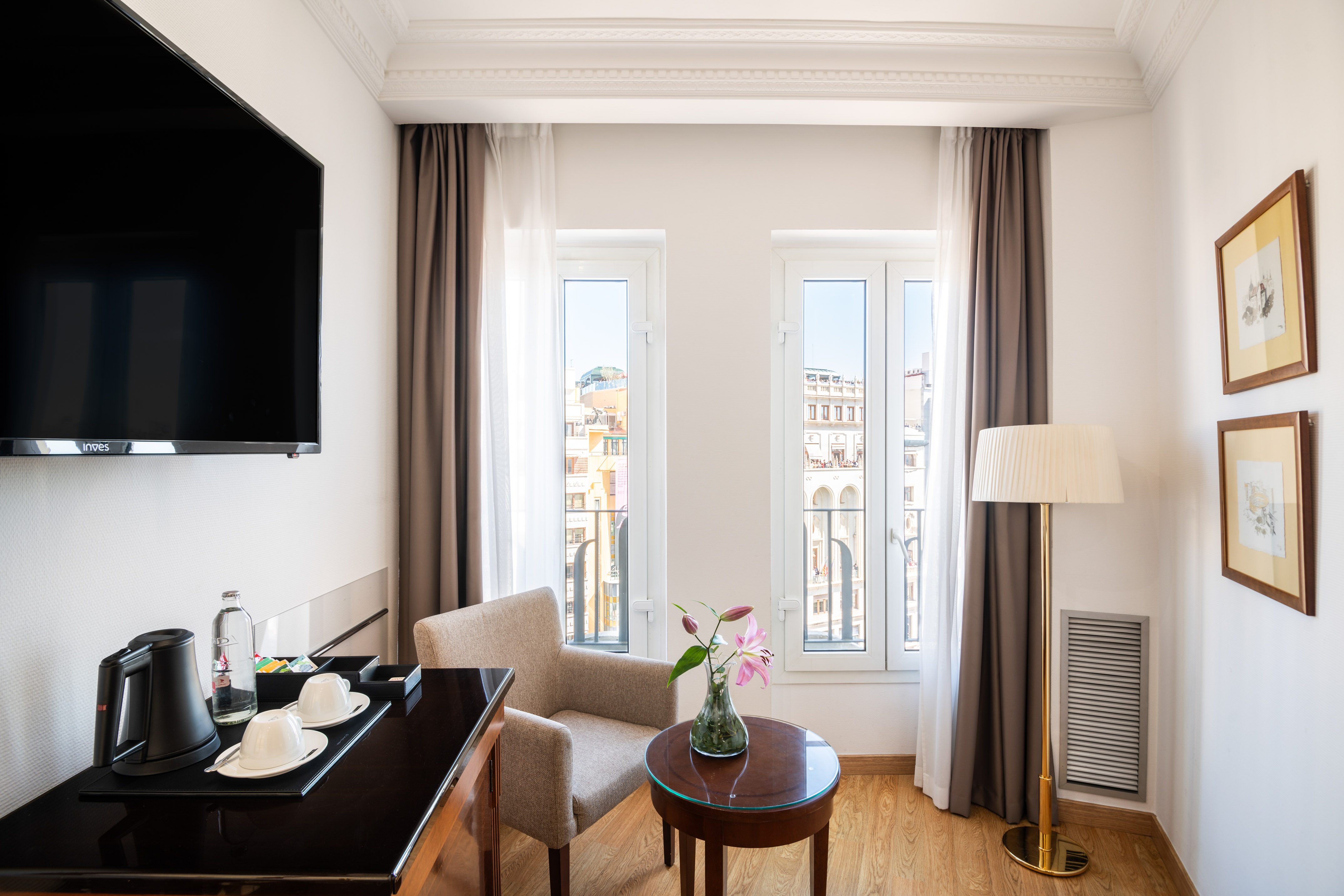 Modern hotel room with TV, coffee station, armchair, and city view windows.