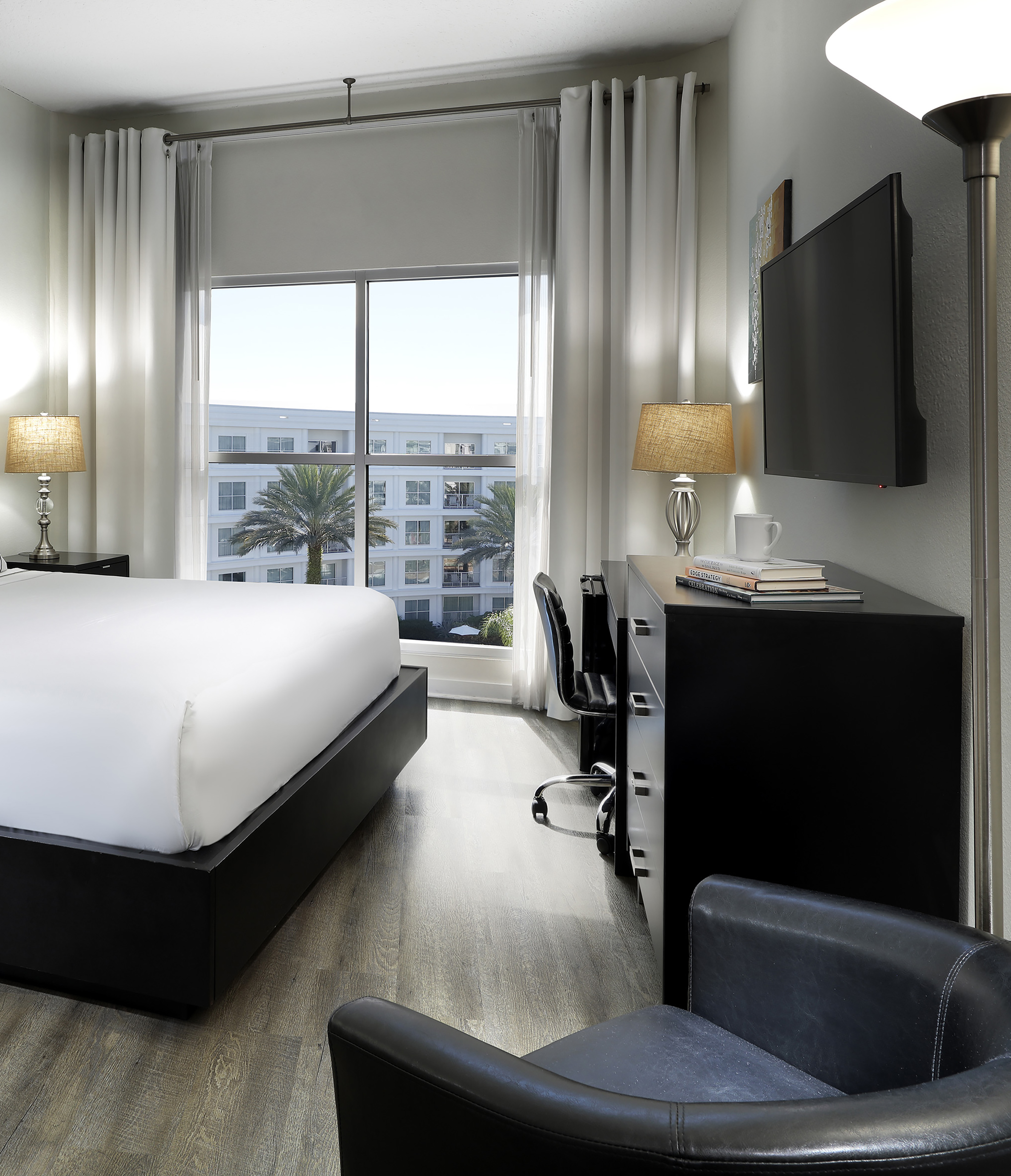 Modern hotel bedroom with king bed, desk, TV, and window view of palm trees.