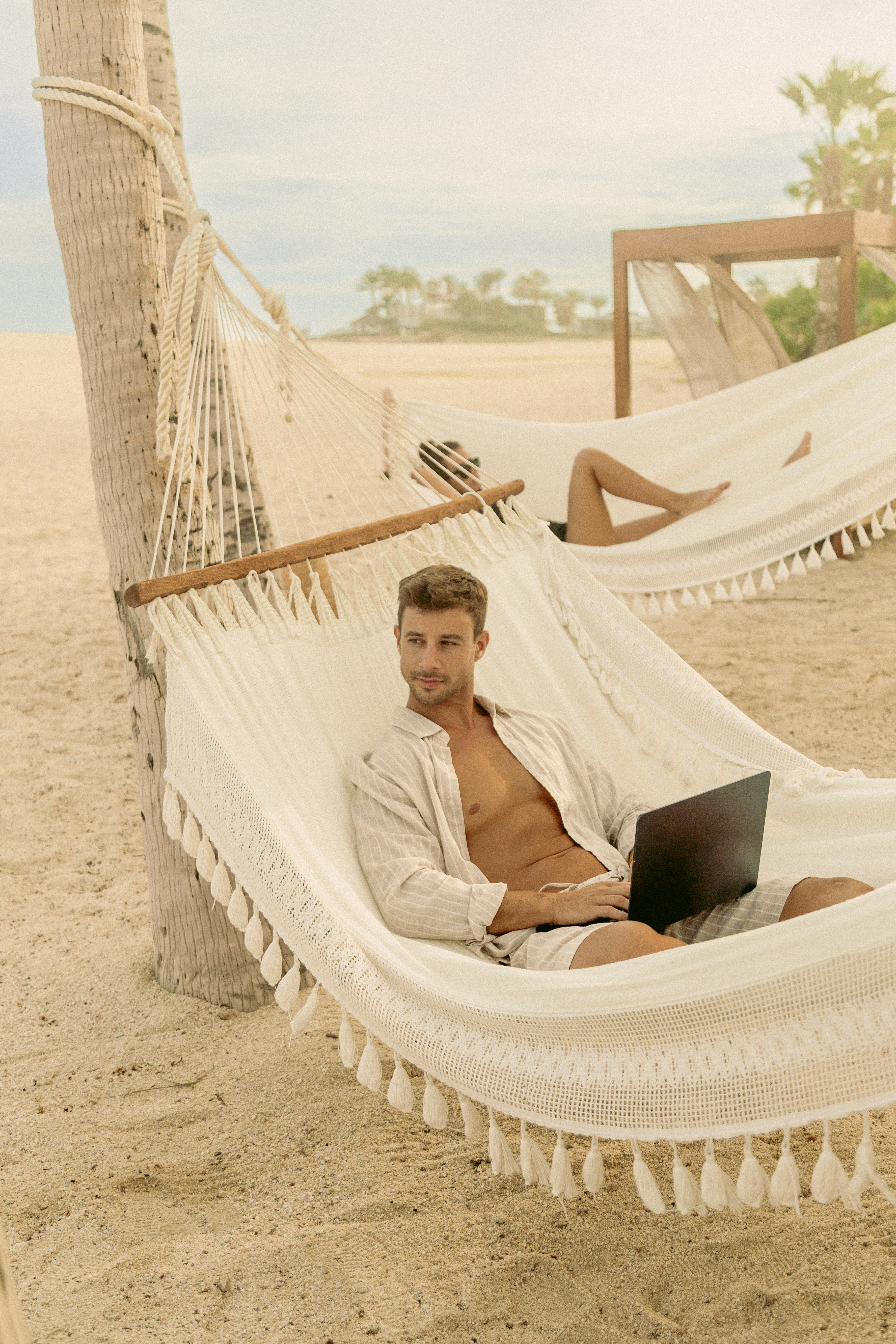 Man working remotely on laptop in white woven hammock on sandy beach.