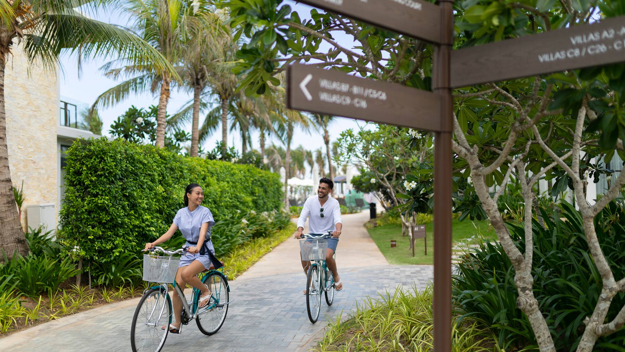 Couple cycling happily on a resort path with palm trees and villa signs.