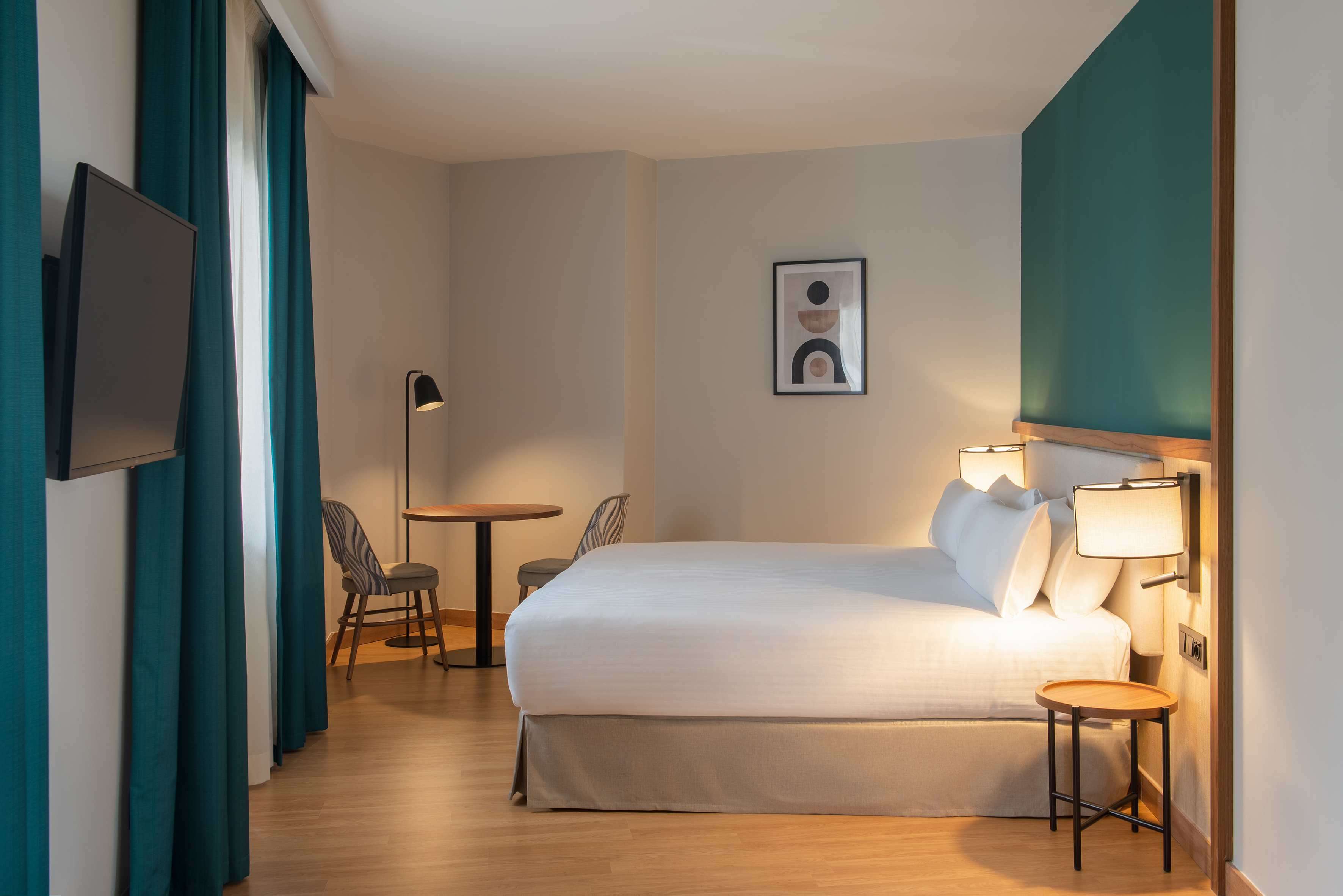 Modern hotel bedroom with white bed, teal accent wall, wall-mounted TV, and small sitting area.