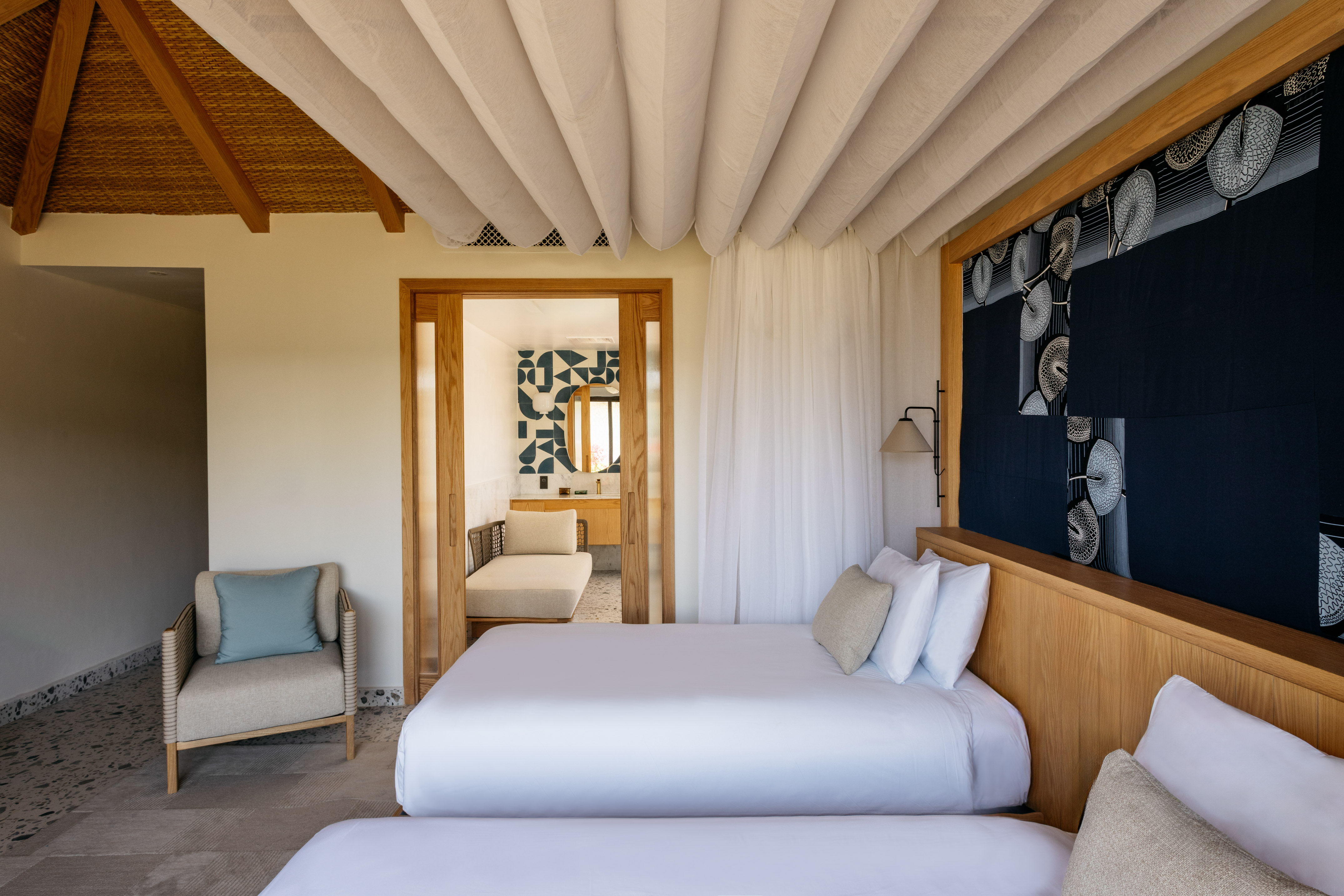 Modern tropical bedroom with two beds, woven ceiling, and open bathroom.