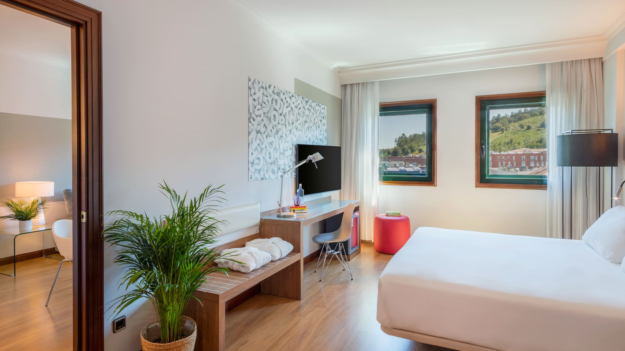 Modern hotel room with white bed, desk, TV, and windows to hills.