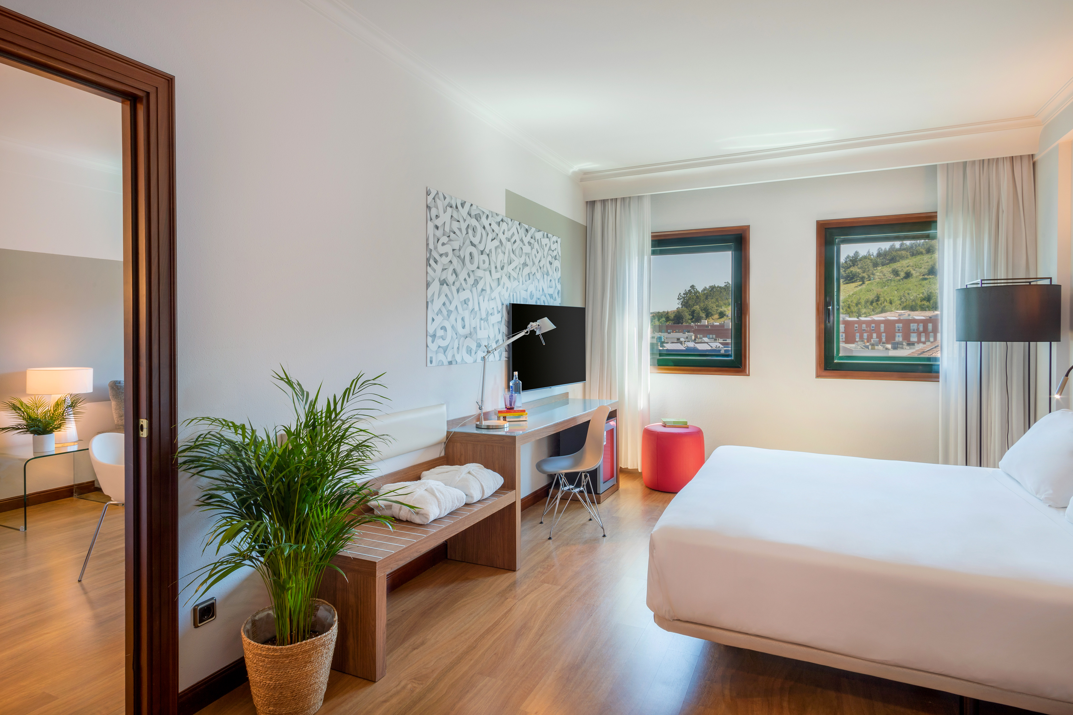 Modern hotel room with white bed, desk, TV, and windows to hills.