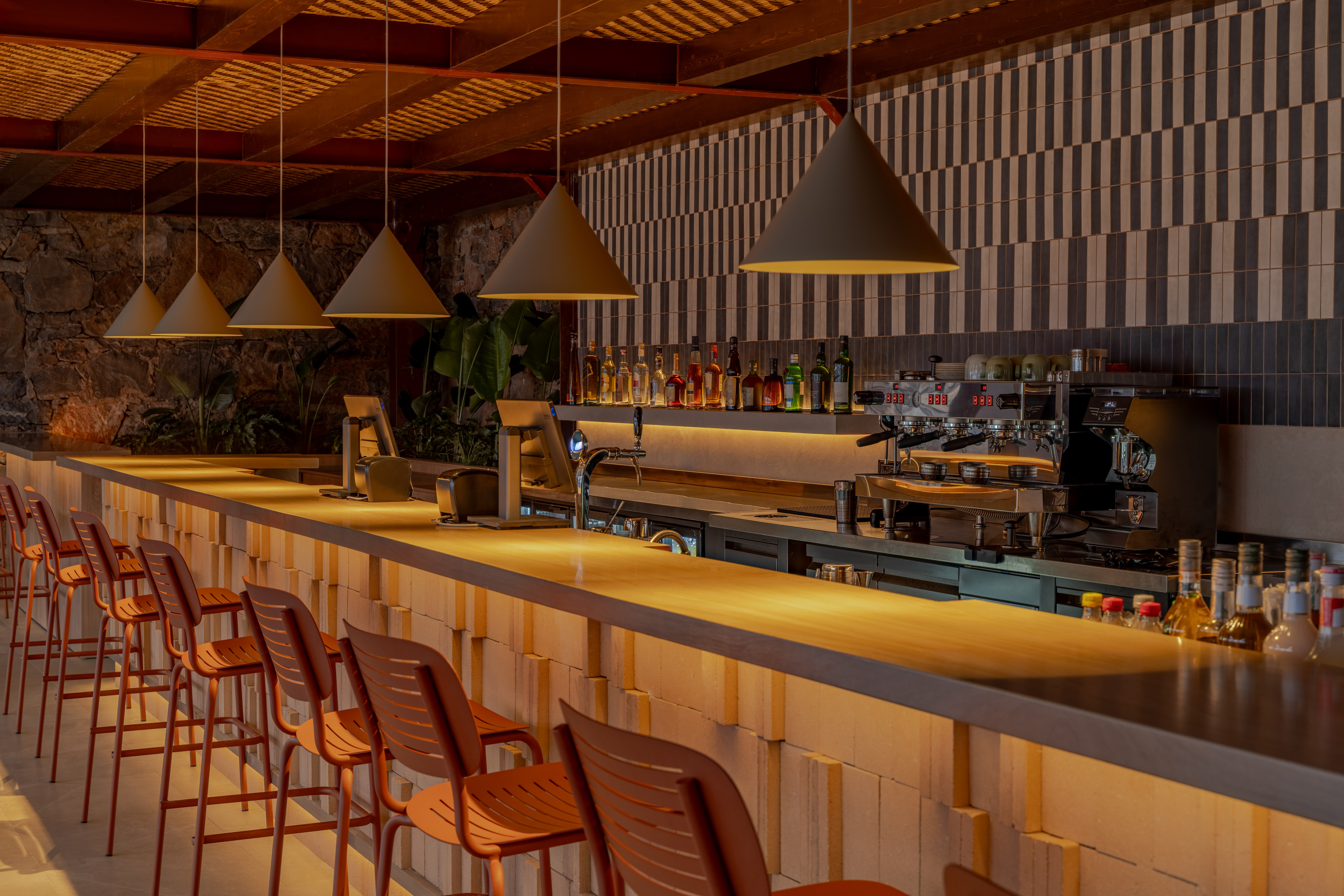 Modern bar counter, orange stools, espresso machine, pendant lights, tiled wall.