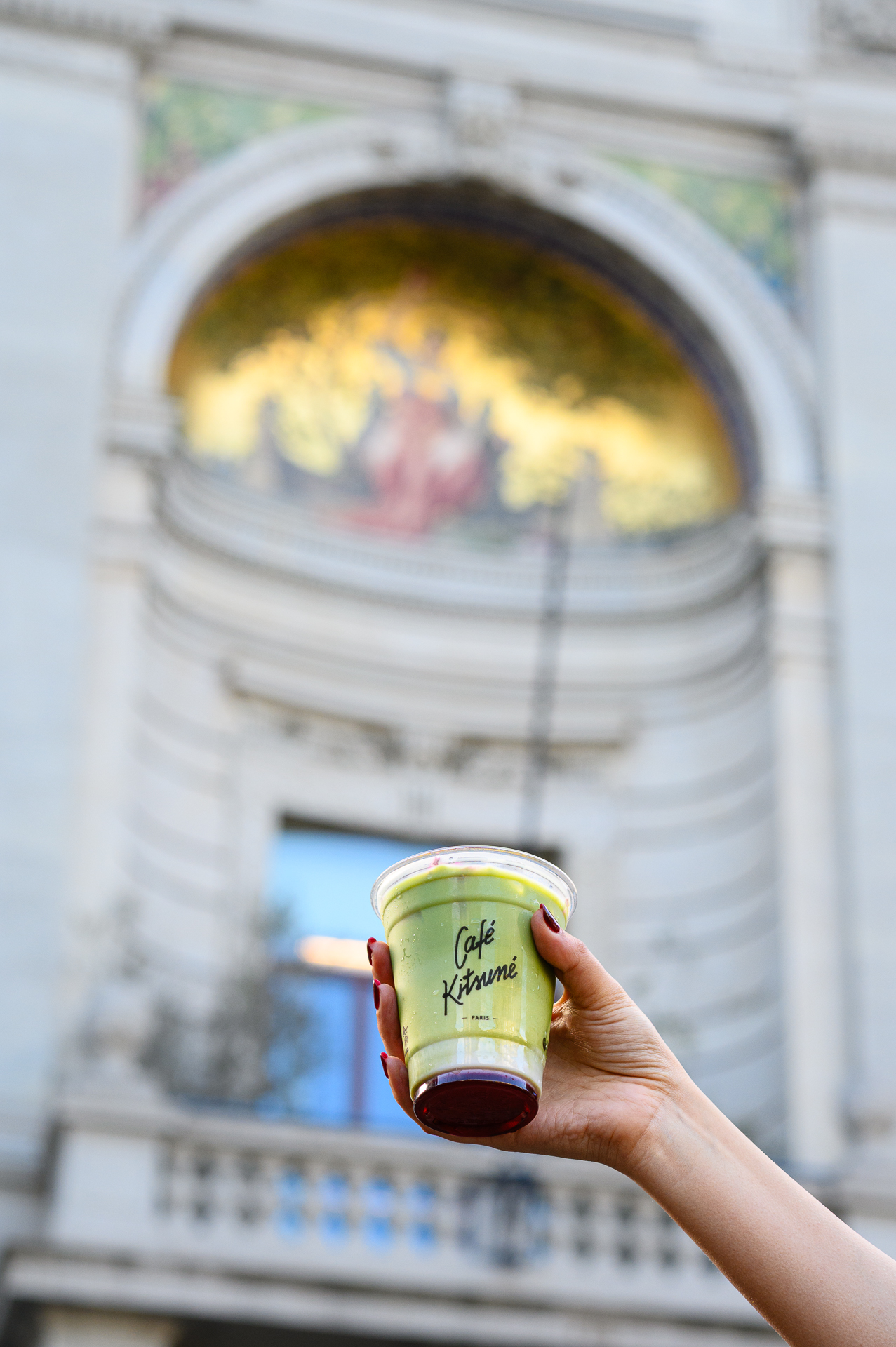 Café Kitsuné matcha latte in hand, blurred mosaic building in Paris background.