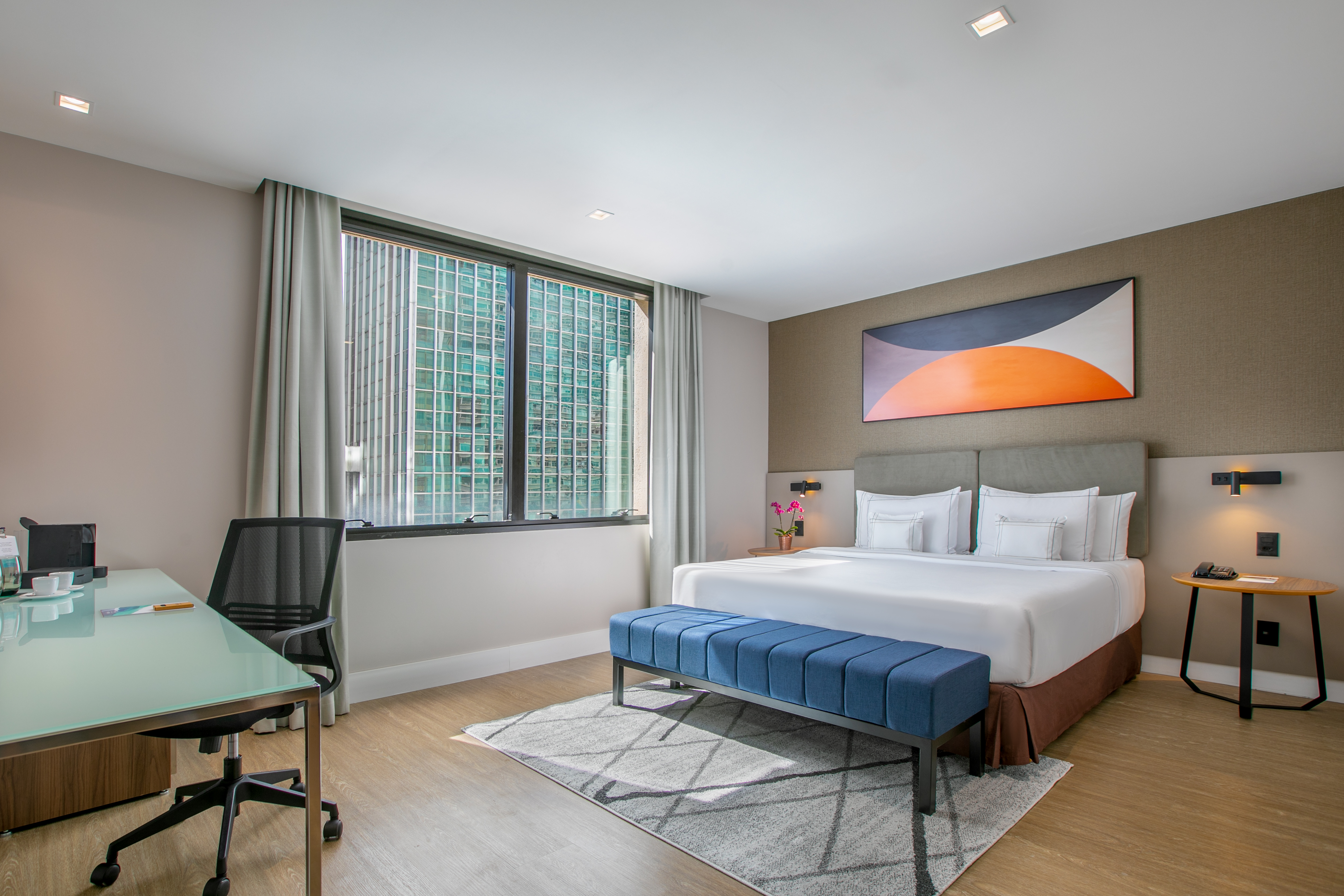 Modern hotel room with king bed, city view window, desk, and abstract art.