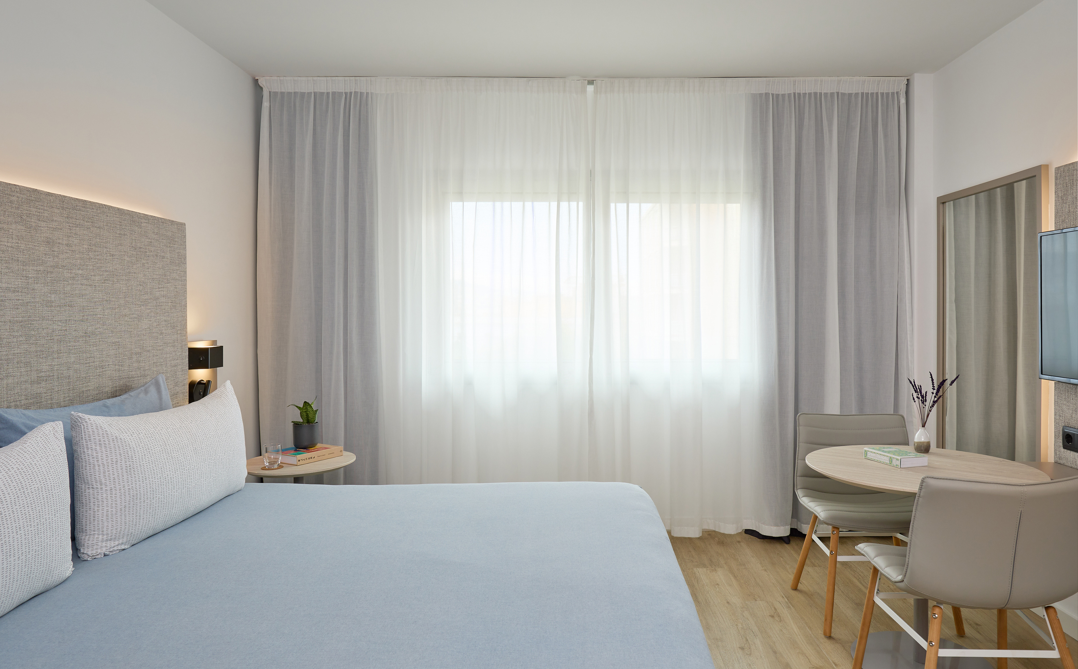 Modern hotel bedroom with light blue bed, large window, and dining area.