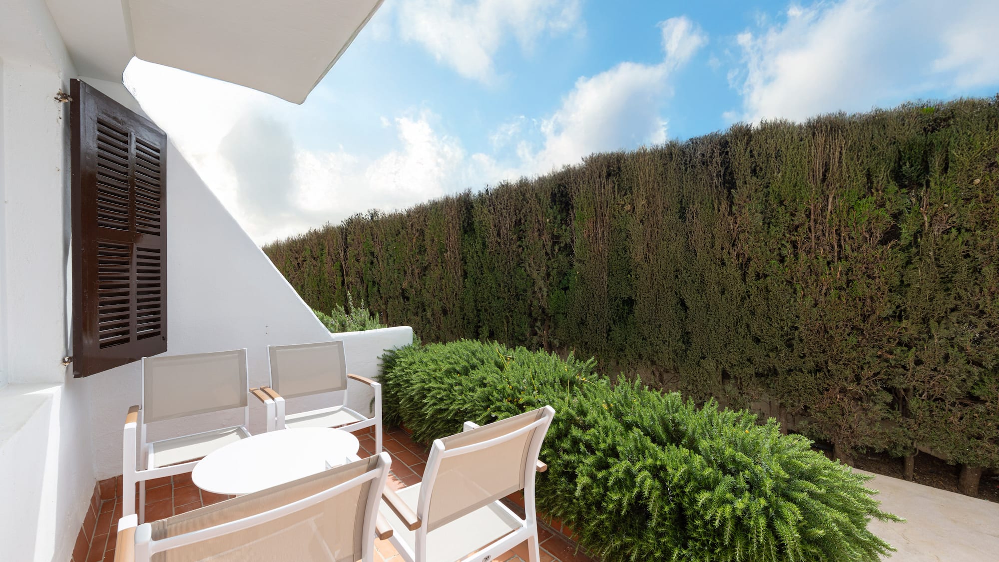 Outdoor terrace with white furniture, terracotta tiles, and green privacy hedge.