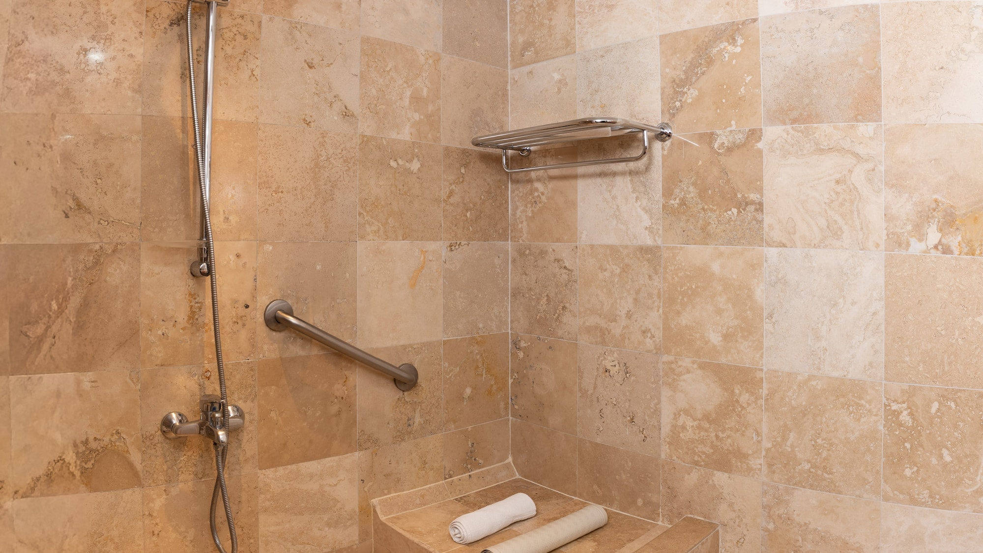 Ducha accesible de travertino, barra de apoyo, asiento y toallas.