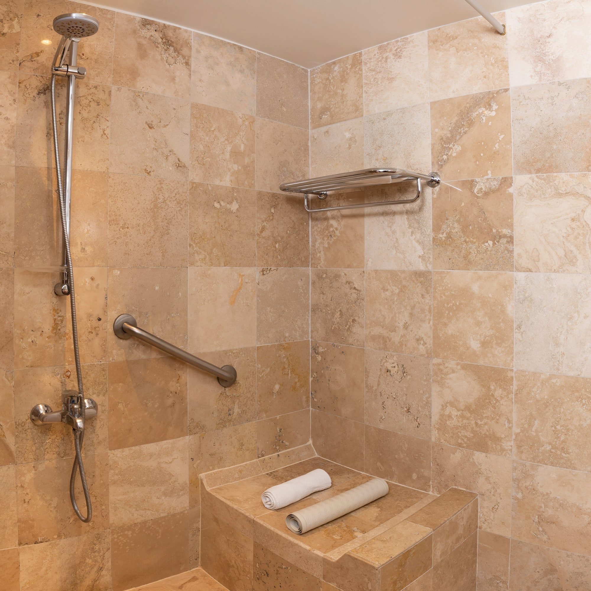 Ducha accesible de travertino, barra de apoyo, asiento y toallas.