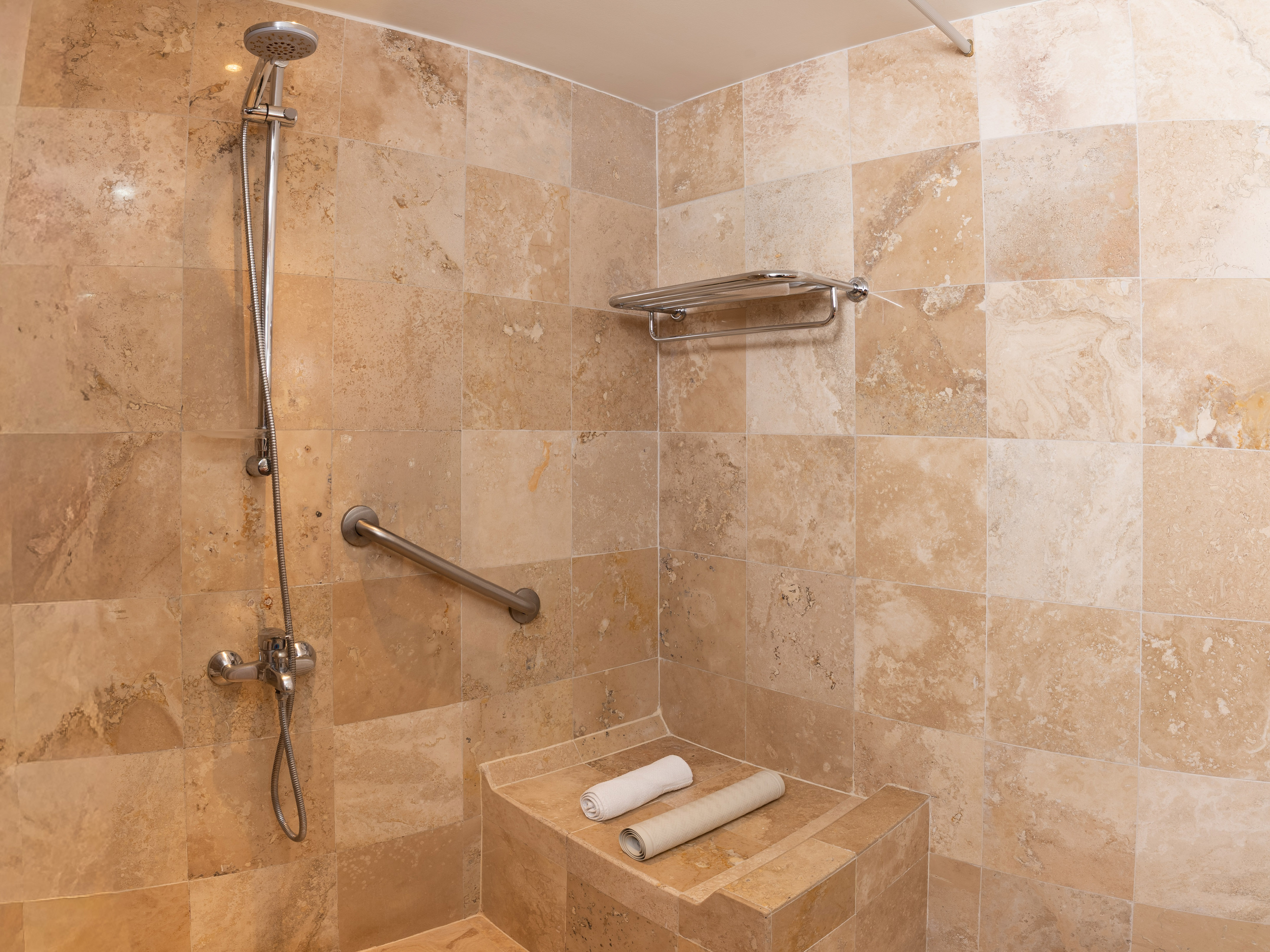 Ducha accesible de travertino, barra de apoyo, asiento y toallas.