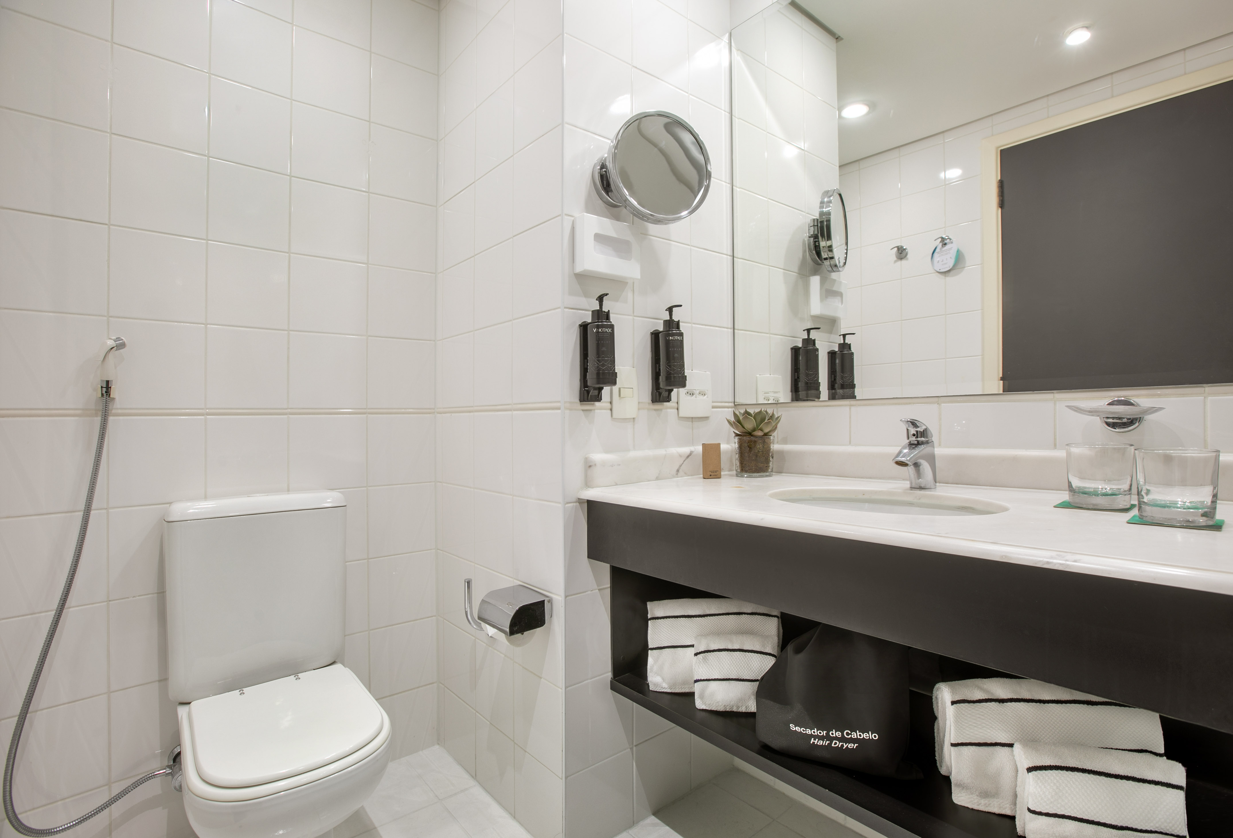 Modern hotel bathroom with white tiles, toilet, sink, mirror, and toiletries.