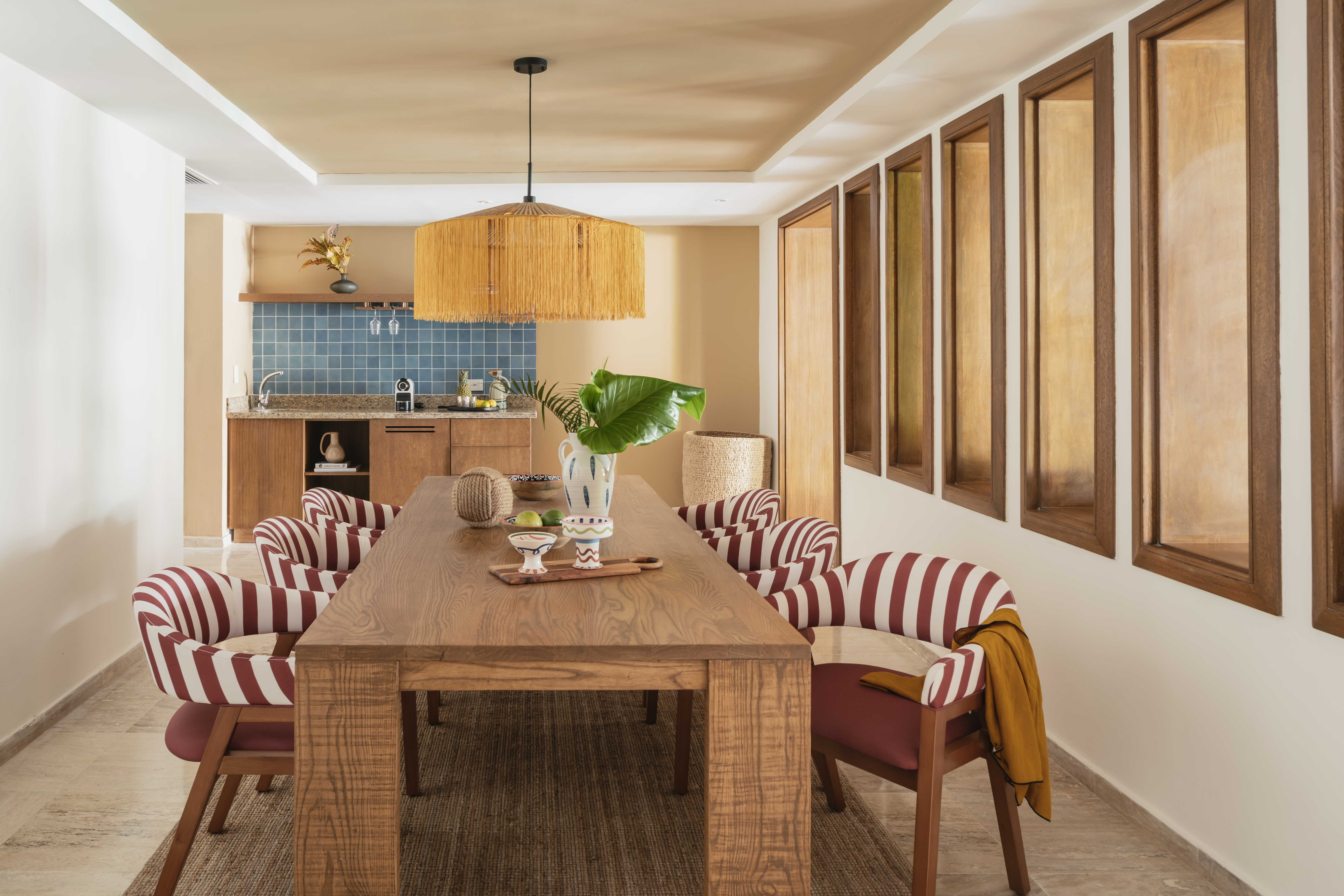 Modern dining room with wood table, red striped chairs, rattan light, kitchenette.