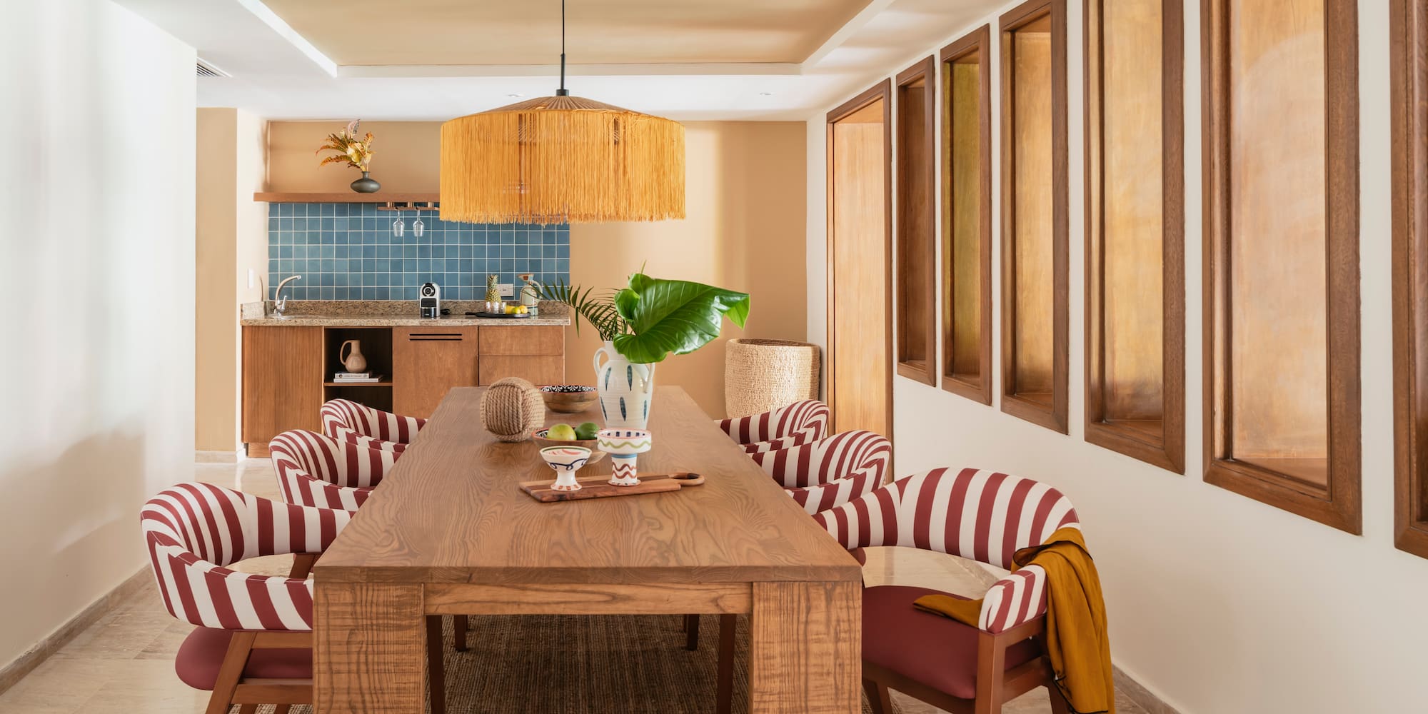 Modern dining room with wood table, red striped chairs, rattan light, kitchenette.