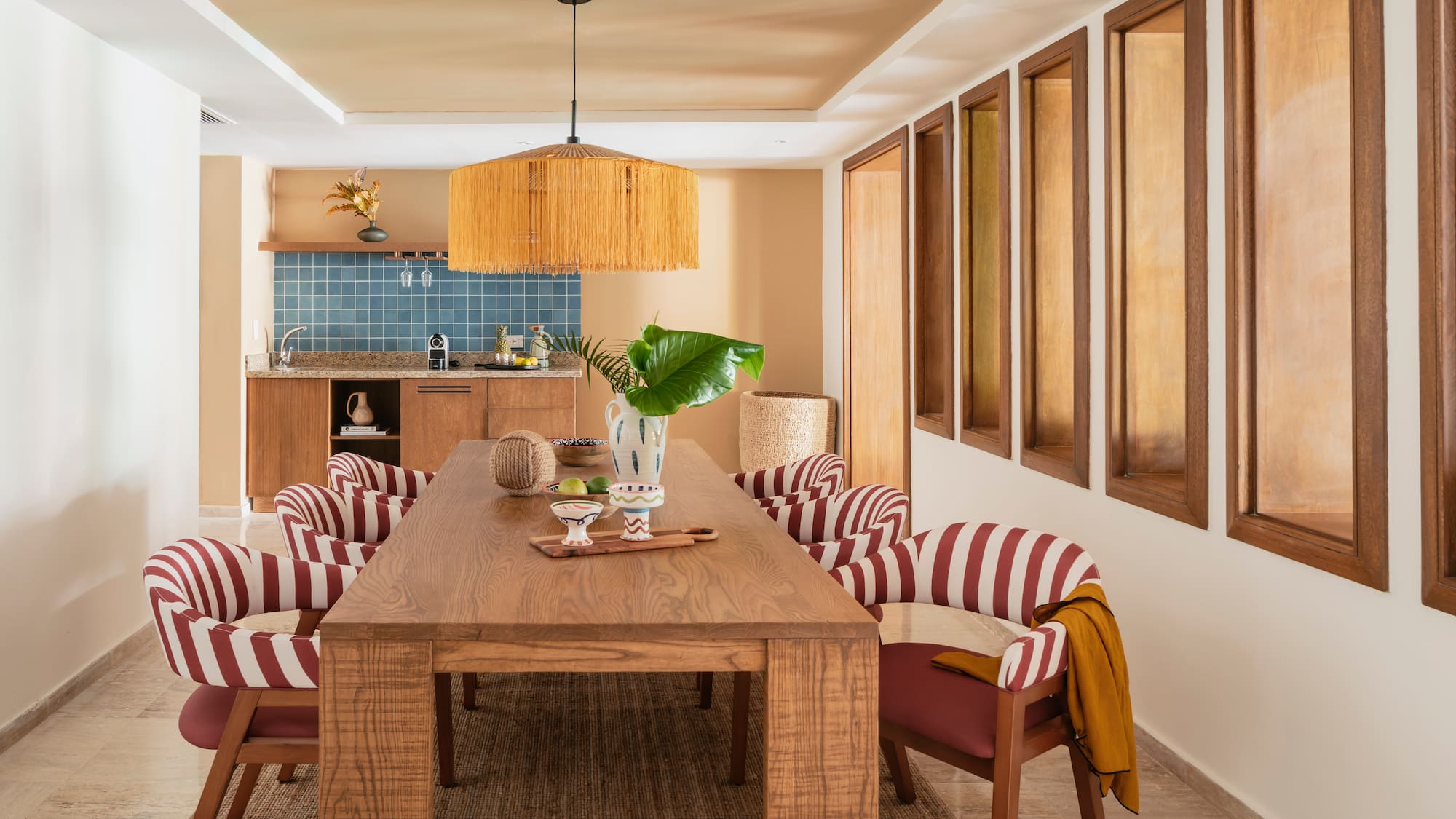 Modern dining room with wood table, red striped chairs, rattan light, kitchenette.