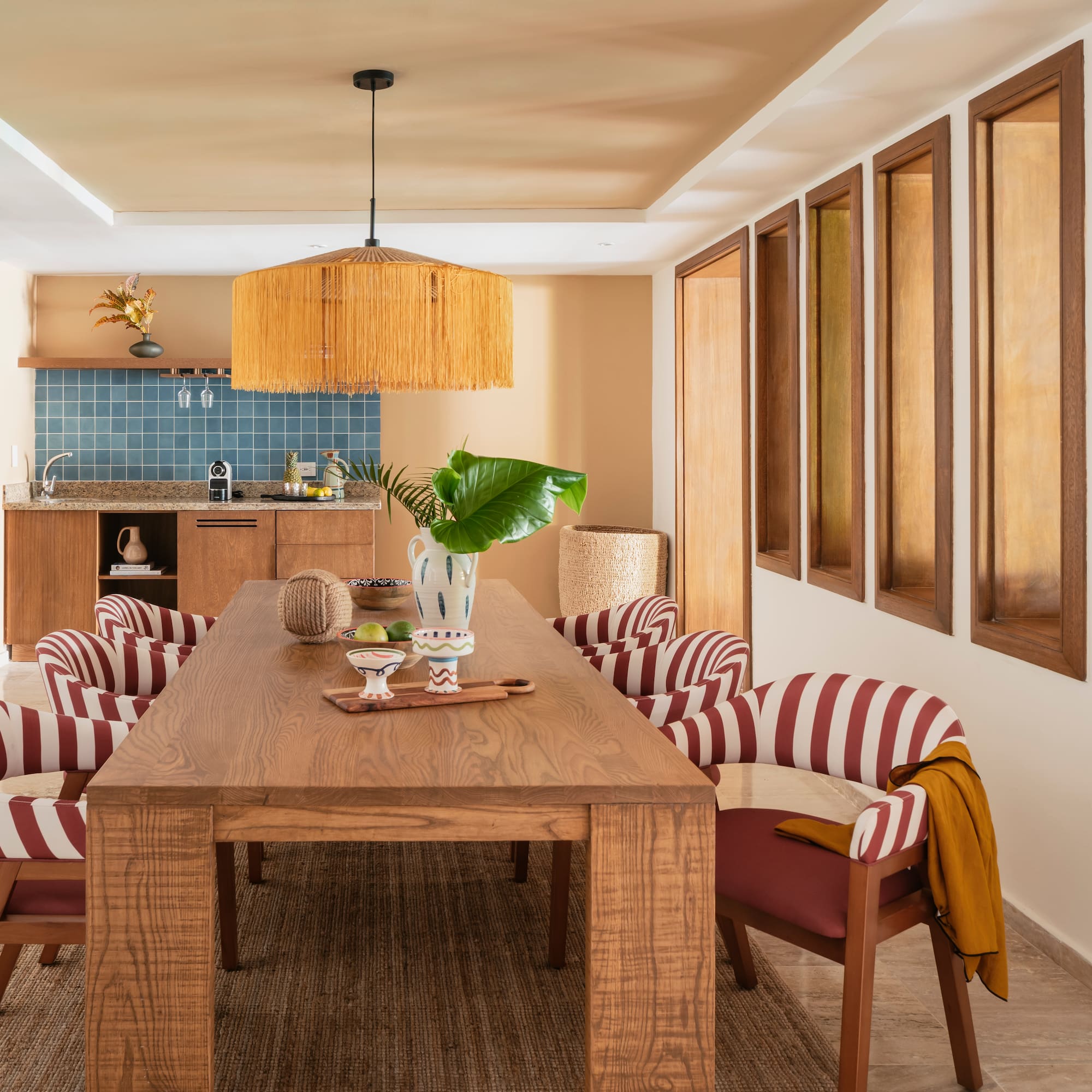 Modern dining room with wood table, red striped chairs, rattan light, kitchenette.