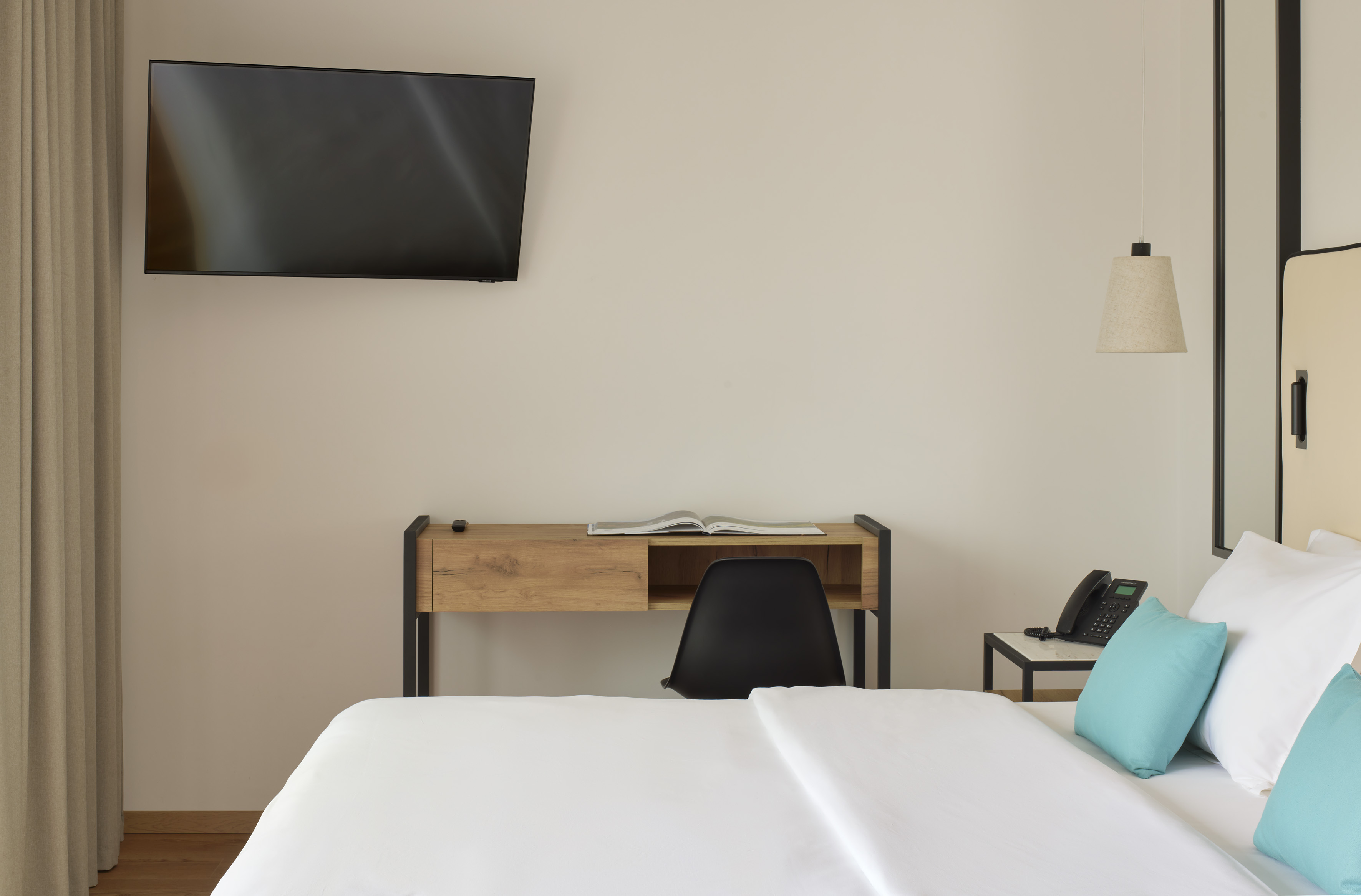 Modern hotel room with TV, work desk, and white bed with teal pillows.