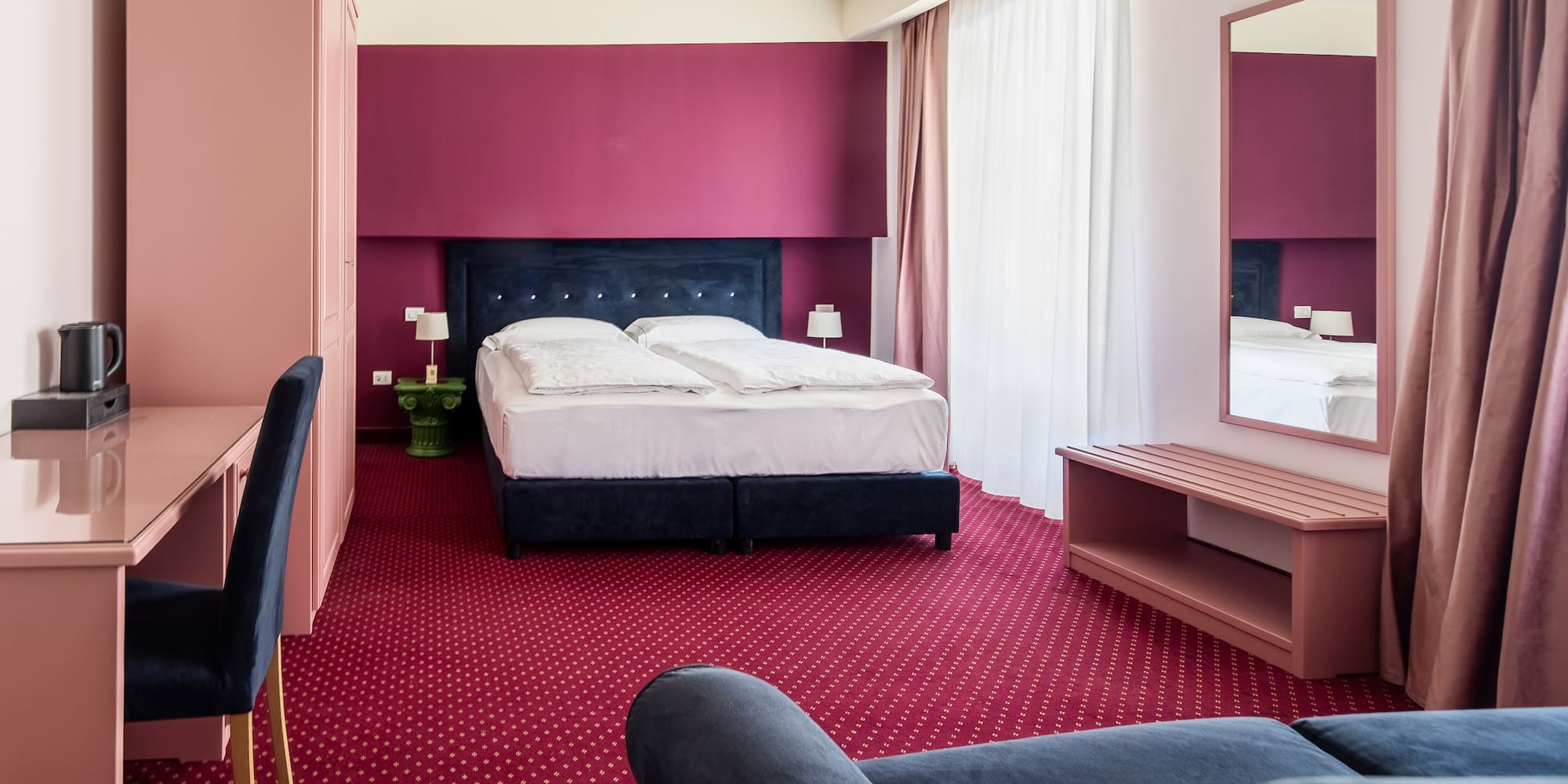 Hotel room with red carpet, pink desk, dark bed, and maroon accent wall.