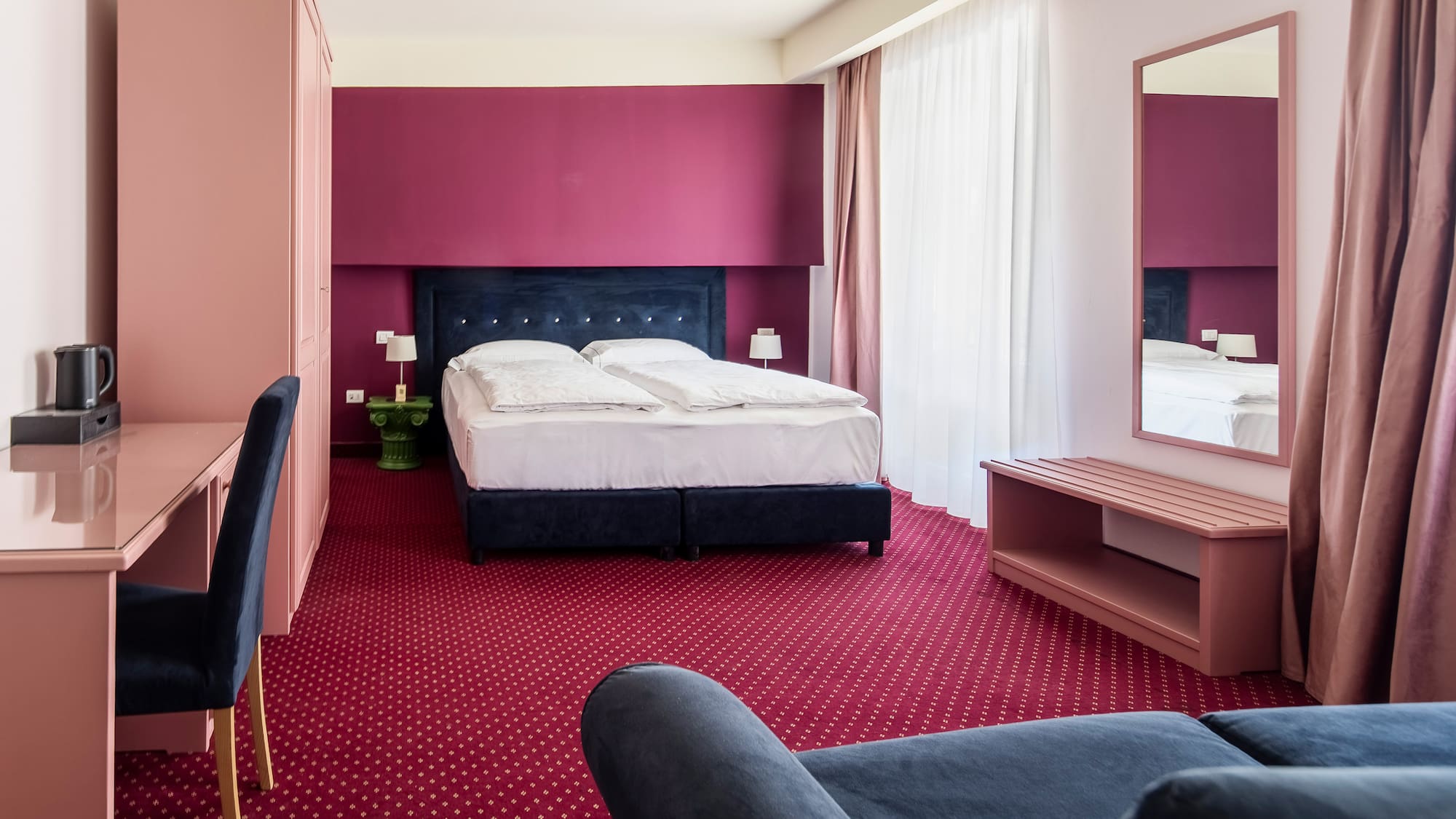 Colorful hotel bedroom with magenta wall, red carpet, blue bed, and pink desk.