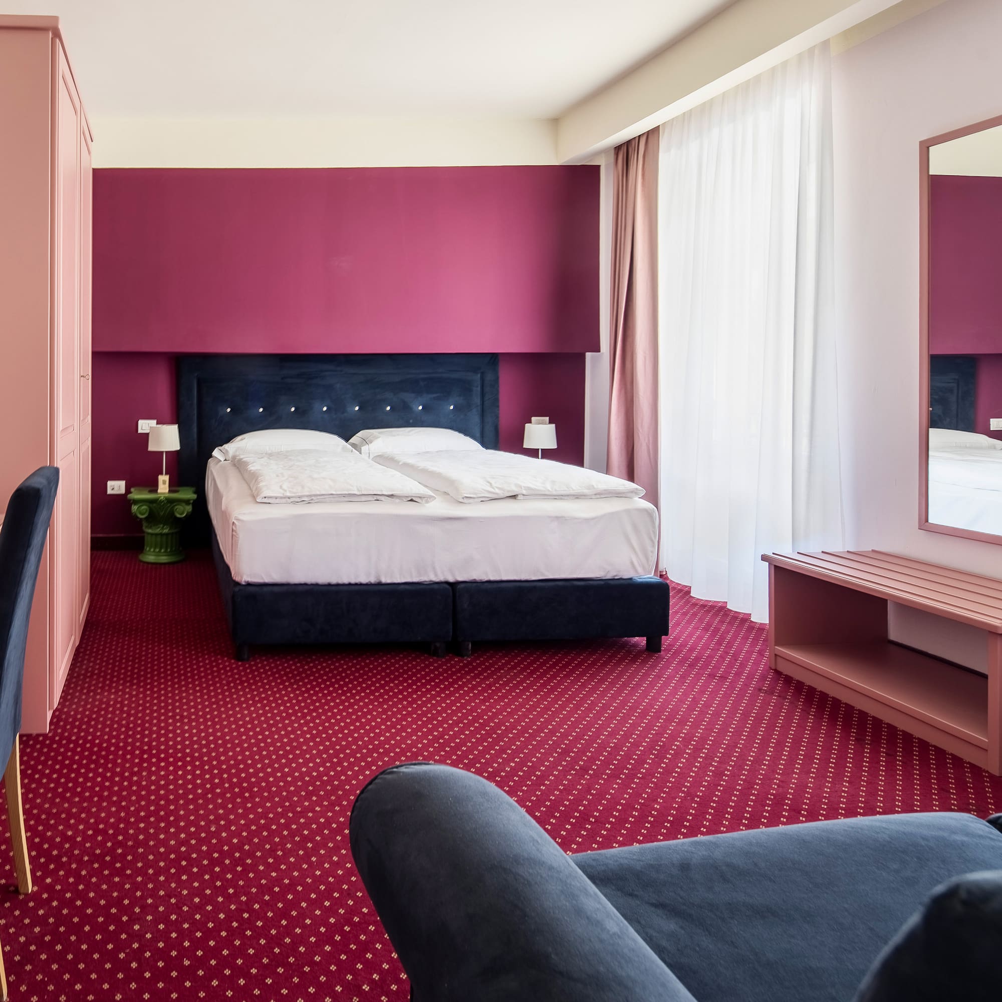 Hotel room with red carpet, pink desk, dark bed, and maroon accent wall.