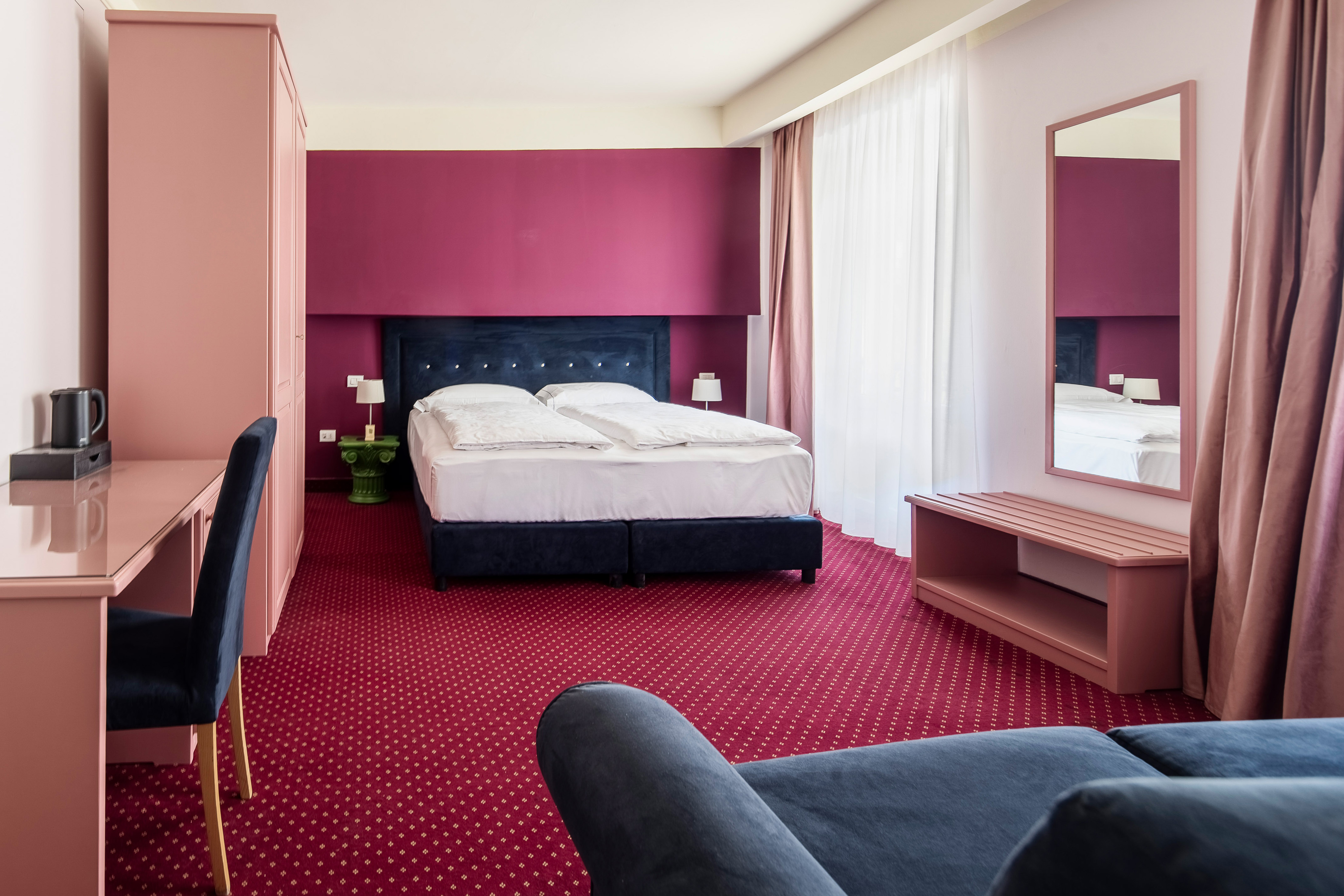 Hotel room with red carpet, pink desk, dark bed, and maroon accent wall.