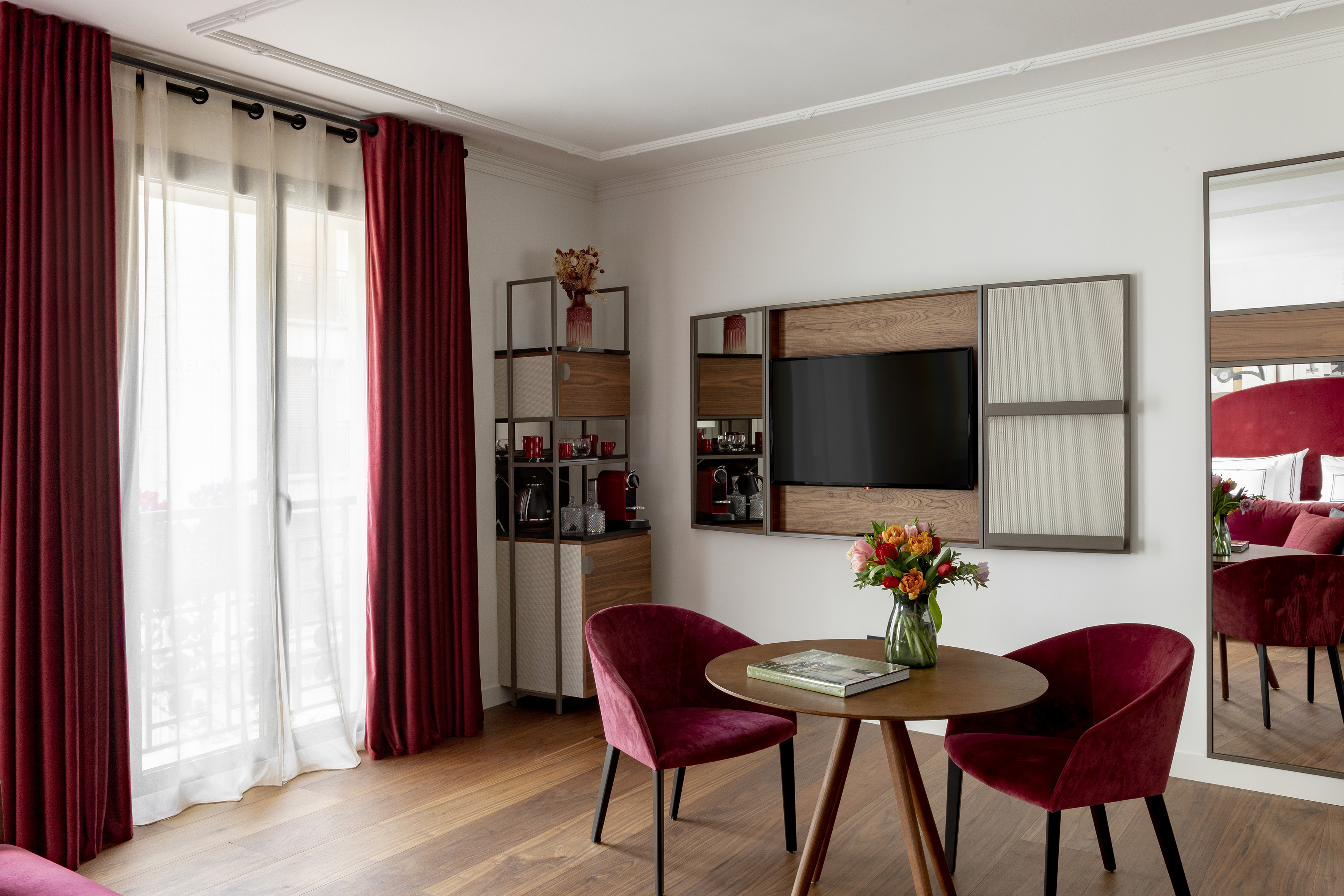 Modern hotel living area with red velvet chairs, minibar, TV, wood table.