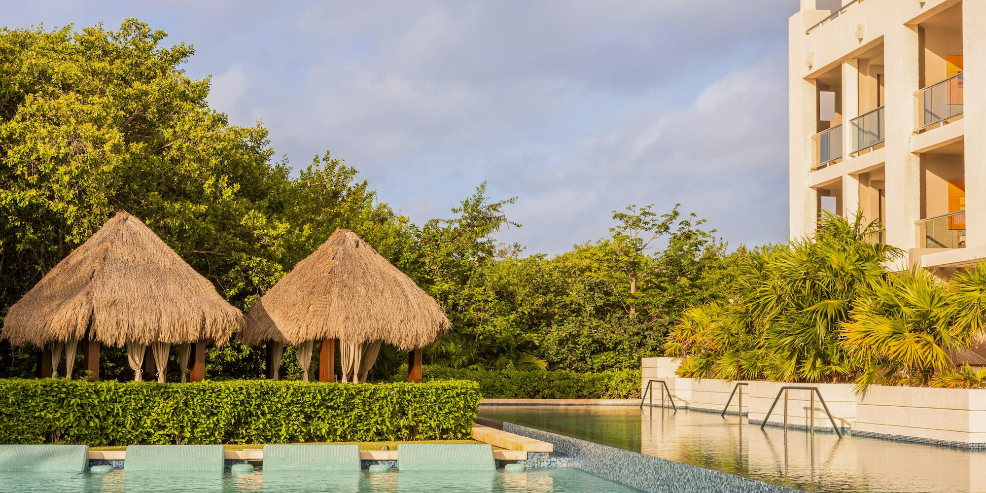 Luxury tropical resort pool, two palapa cabanas, and hotel building.