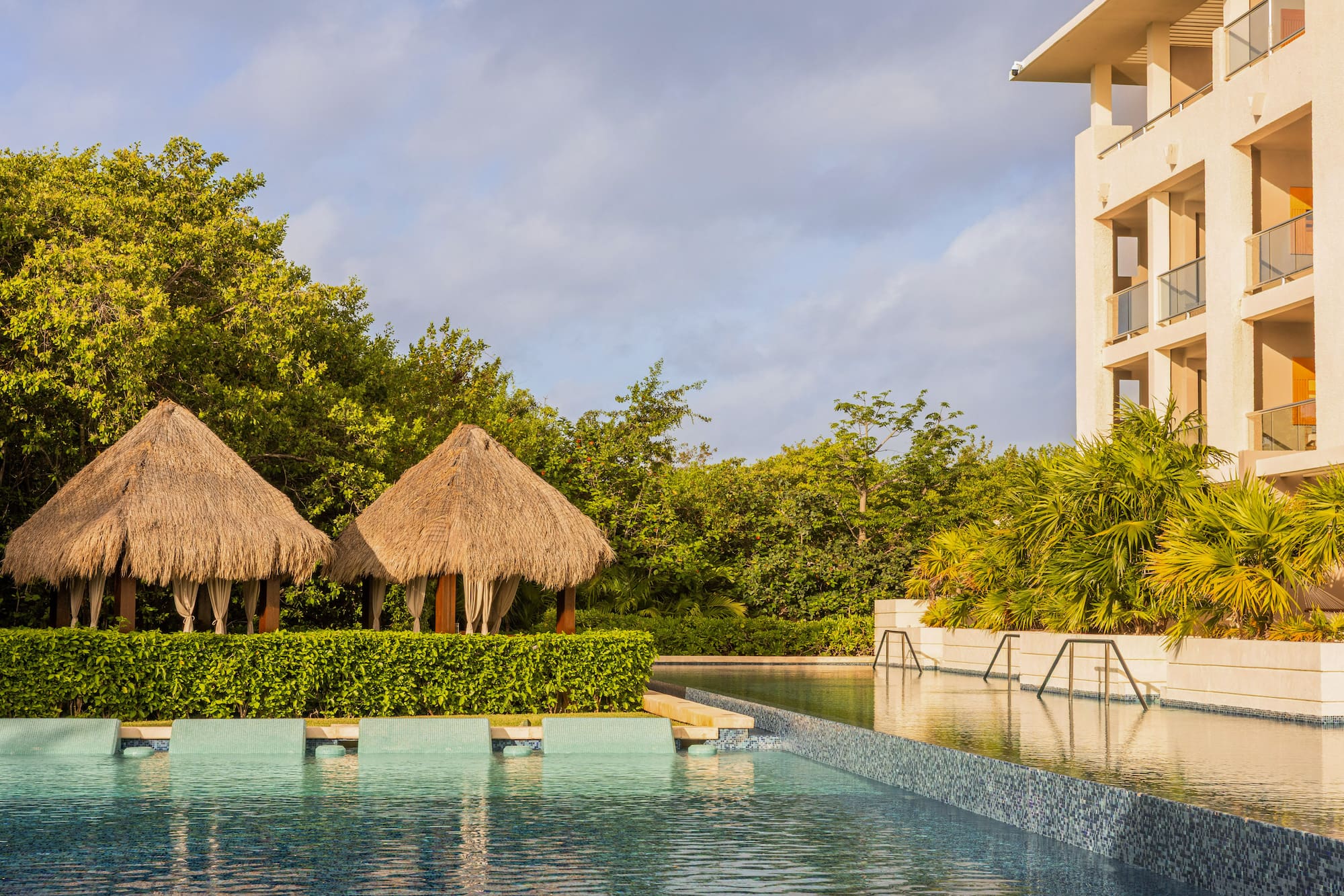 Luxury tropical resort pool, two palapa cabanas, and hotel building.
