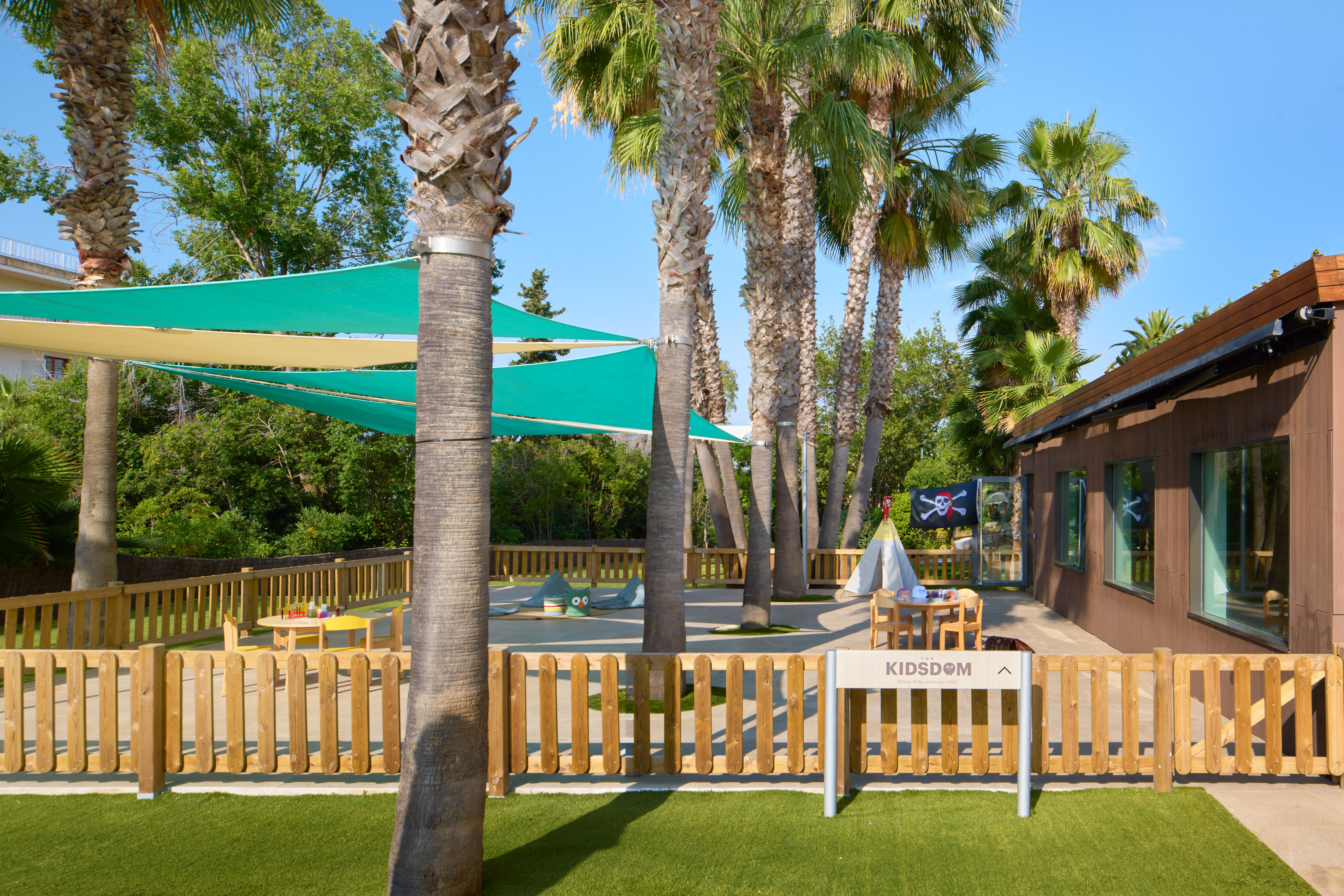 Kidsdom outdoor playground with palm trees, green shade sails, and pirate flag.