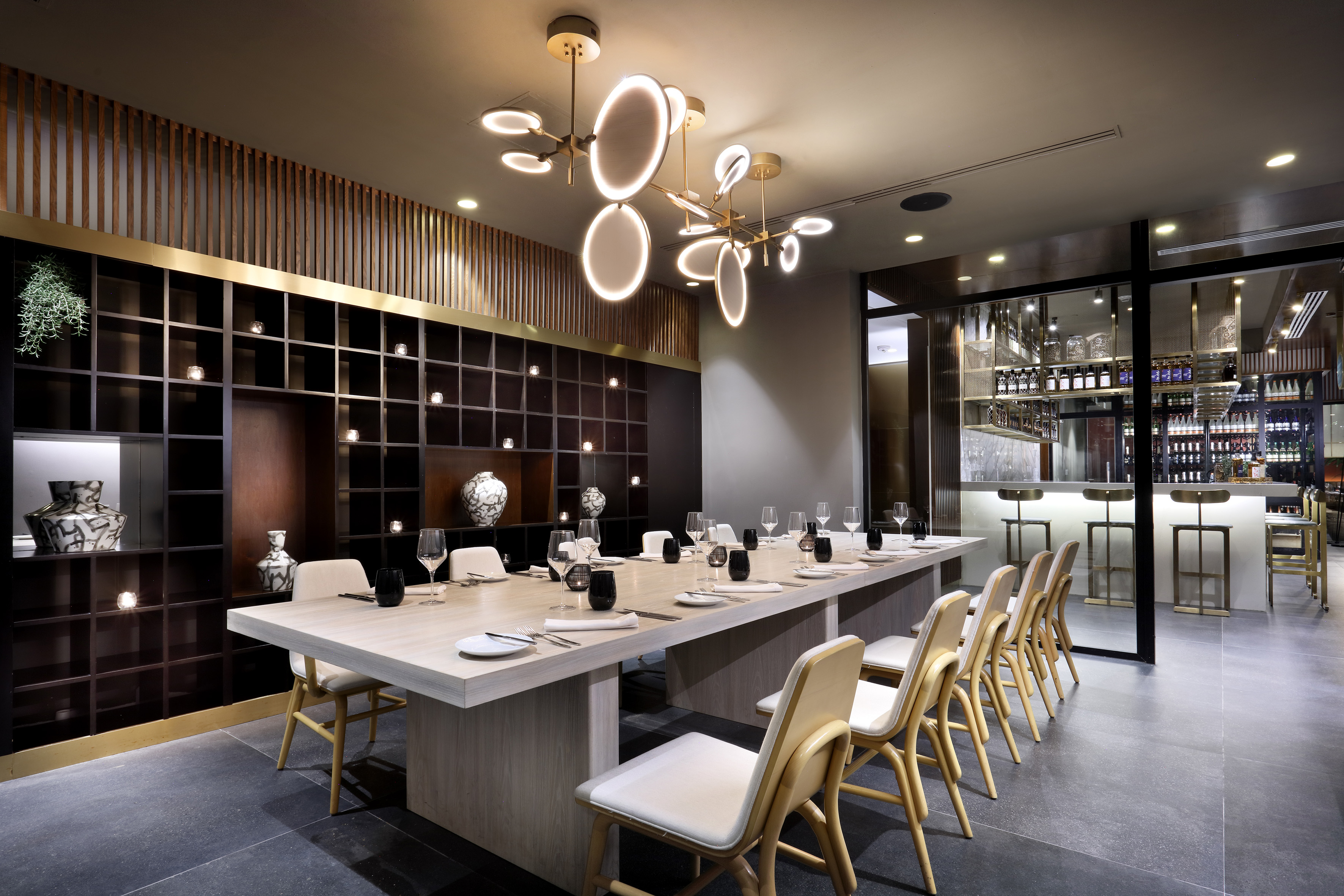 Modern private dining room with long table, unique chandeliers, and adjacent bar.
