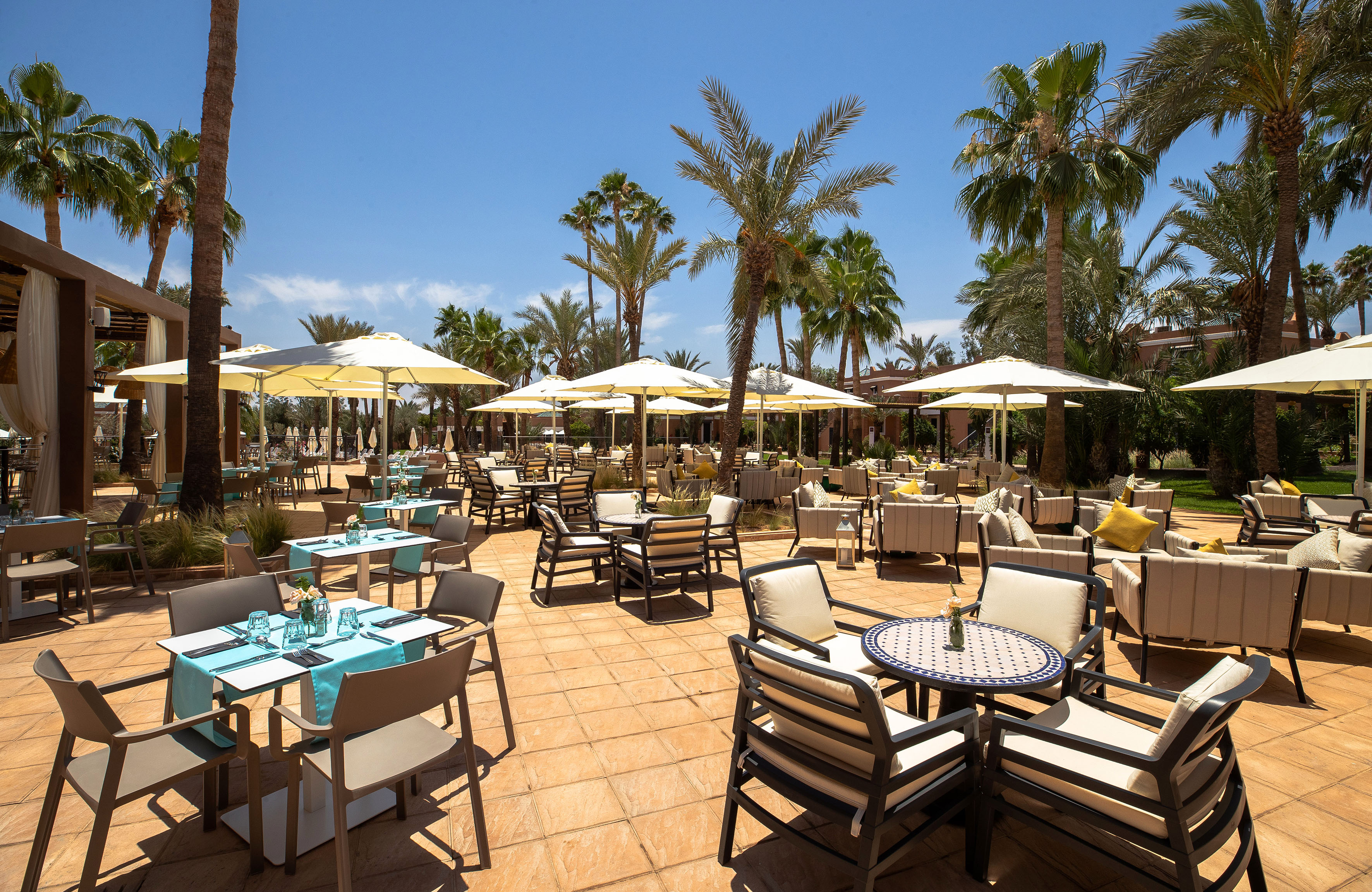 Sunny outdoor restaurant patio with tables, cream umbrellas, and palm trees.